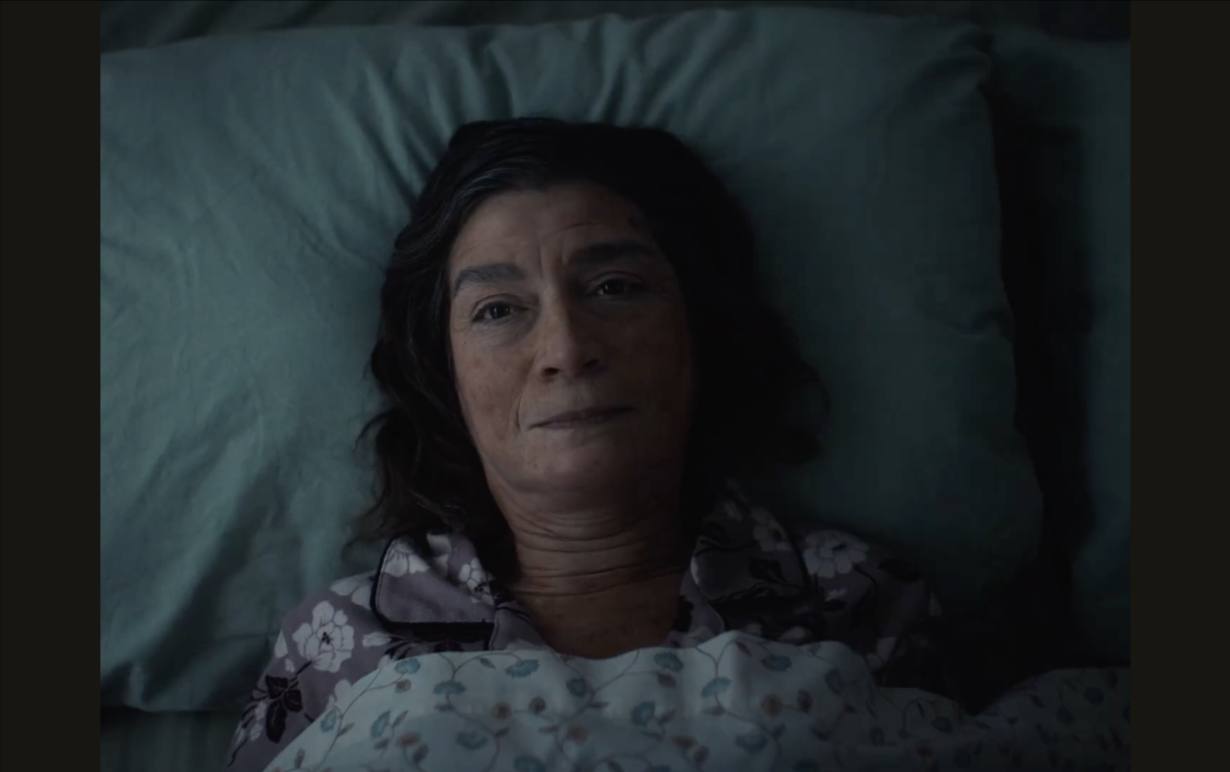 A woman with dark hair lying in bed, looking at the camera, wearing a floral-patterned shirt, with a pillow and teal bedsheet visible.