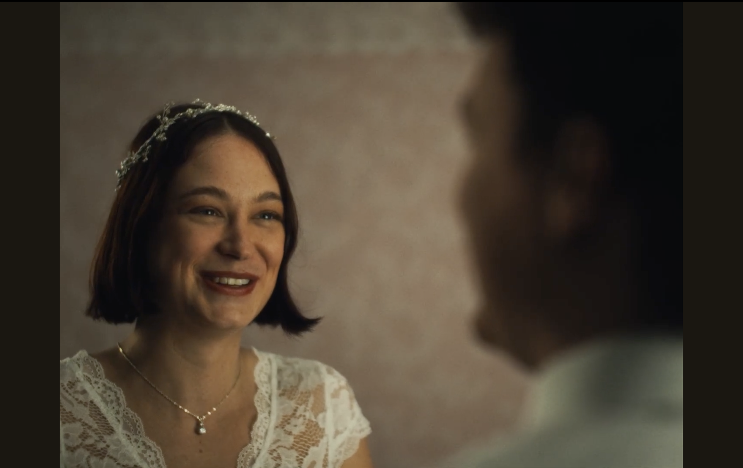 A woman with short brown hair wearing a pearl headband, lace white top, and jewelry, smiling at someone who's visible only partially.