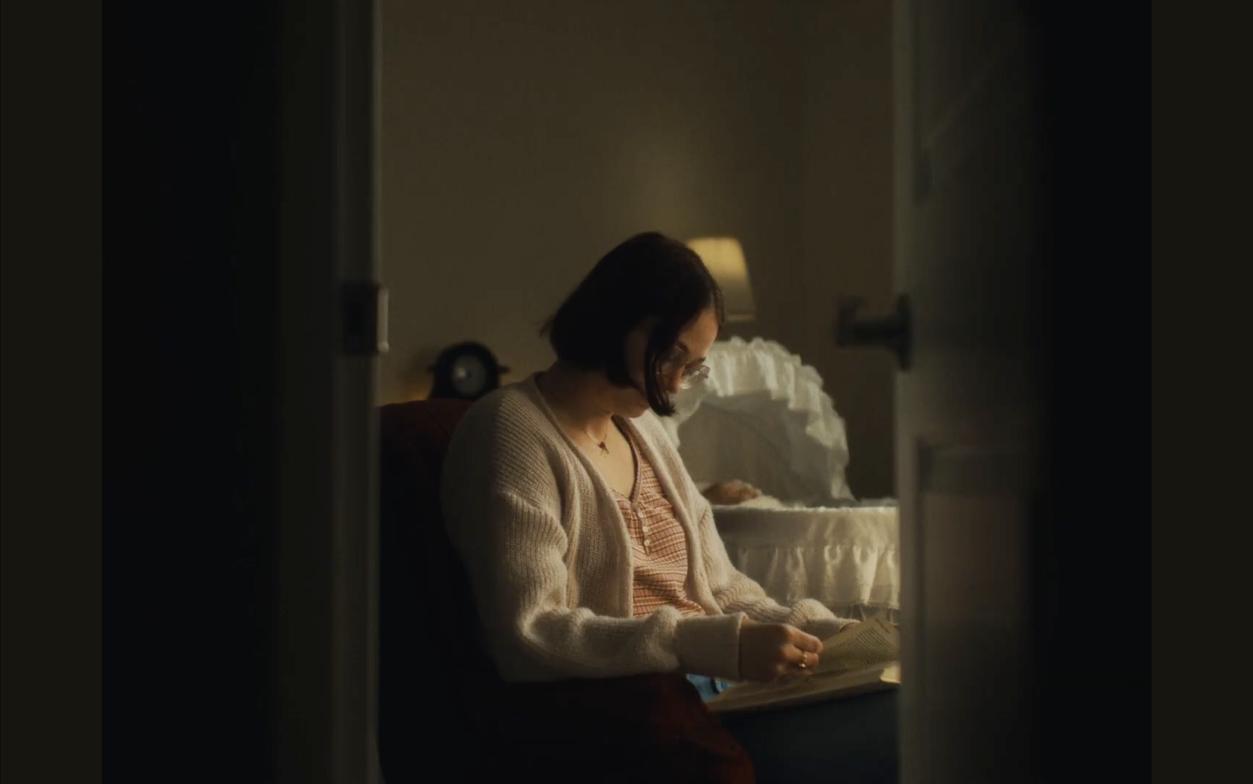A woman with glasses and dark hair sits on a bed, reading a book. The room is softly lit with a warm lamp in the background.
