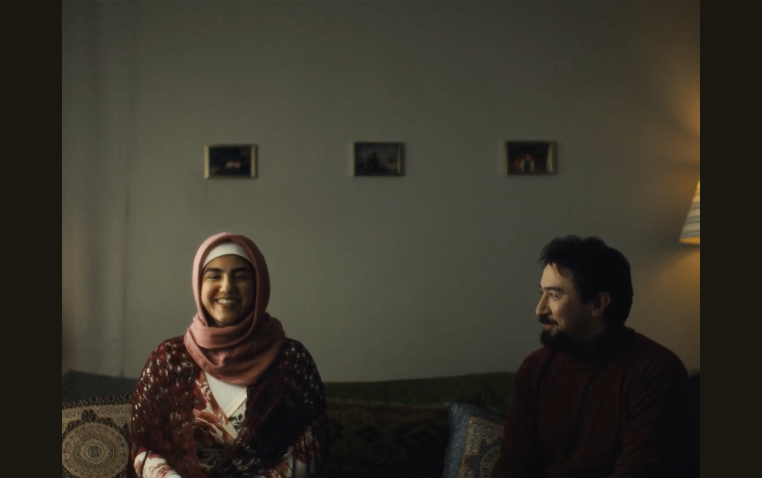 Smiling woman wearing a headscarf, sitting on a patterned cushion, and a man with dark hair and a beard sitting beside her, in a cozy room with a gray wall, small framed pictures, and warm lighting.