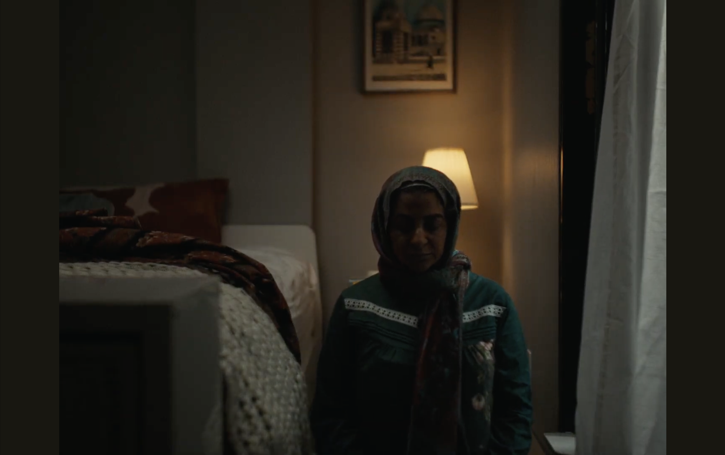 A woman with a headscarf sitting in a dimly lit room near a window with a curtain and a lamp on a side table, background includes a bed and picture on the wall