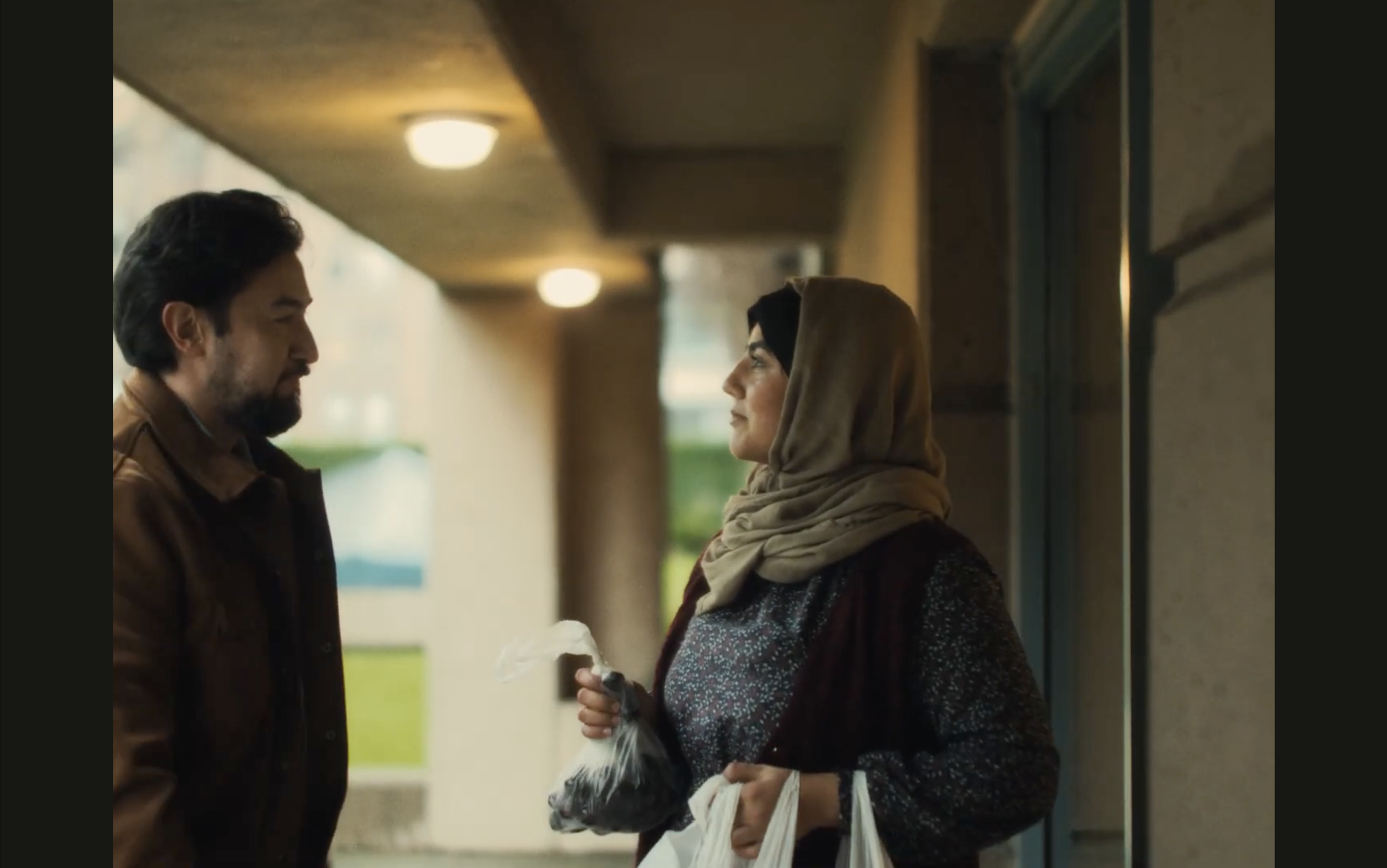 A man and a woman standing face to face on a dimly lit outdoor corridor, with the man holding a white bag and the woman holding a bag of groceries, both making eye contact.
