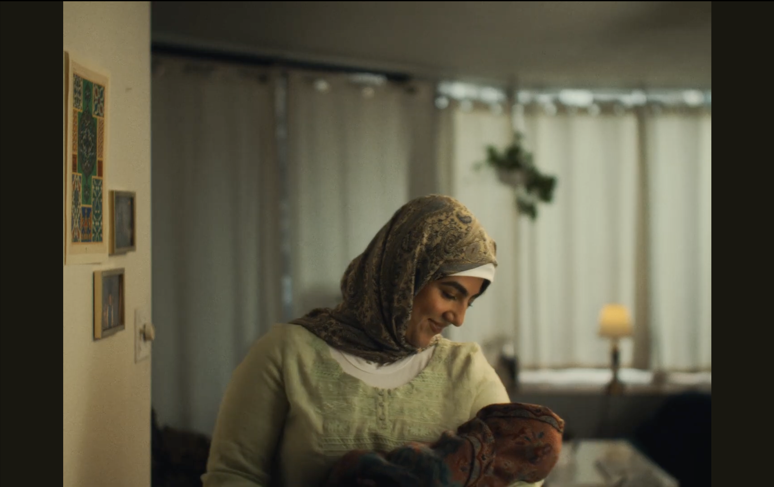 A woman wearing a patterned headscarf and light green top smiling while holding a baby in a cozy living room with curtains and wall art.