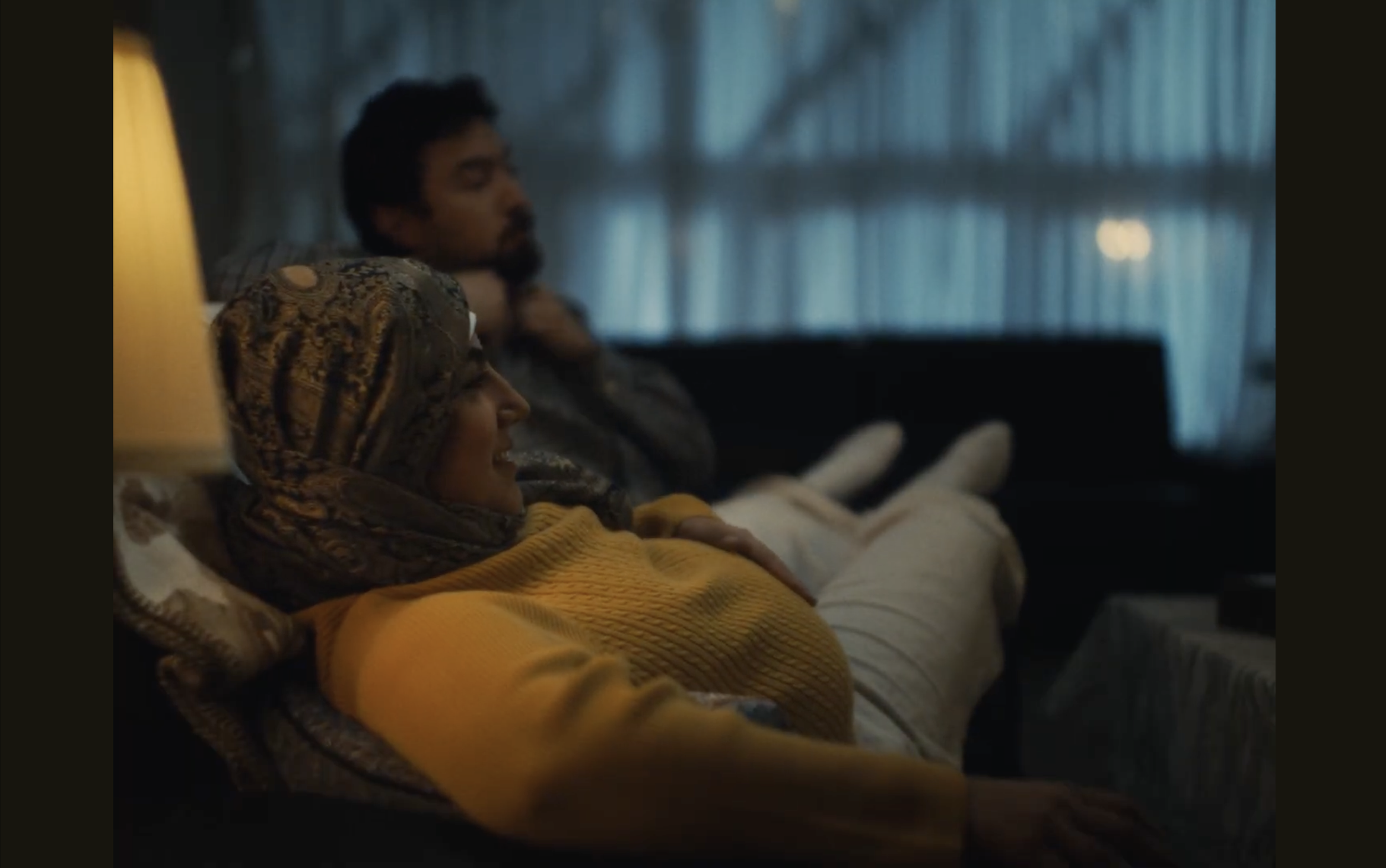 A woman with a headscarf and a man with short hair and a beard sit on a bed in a dimly lit room with blue curtains, relaxed and comfortable.