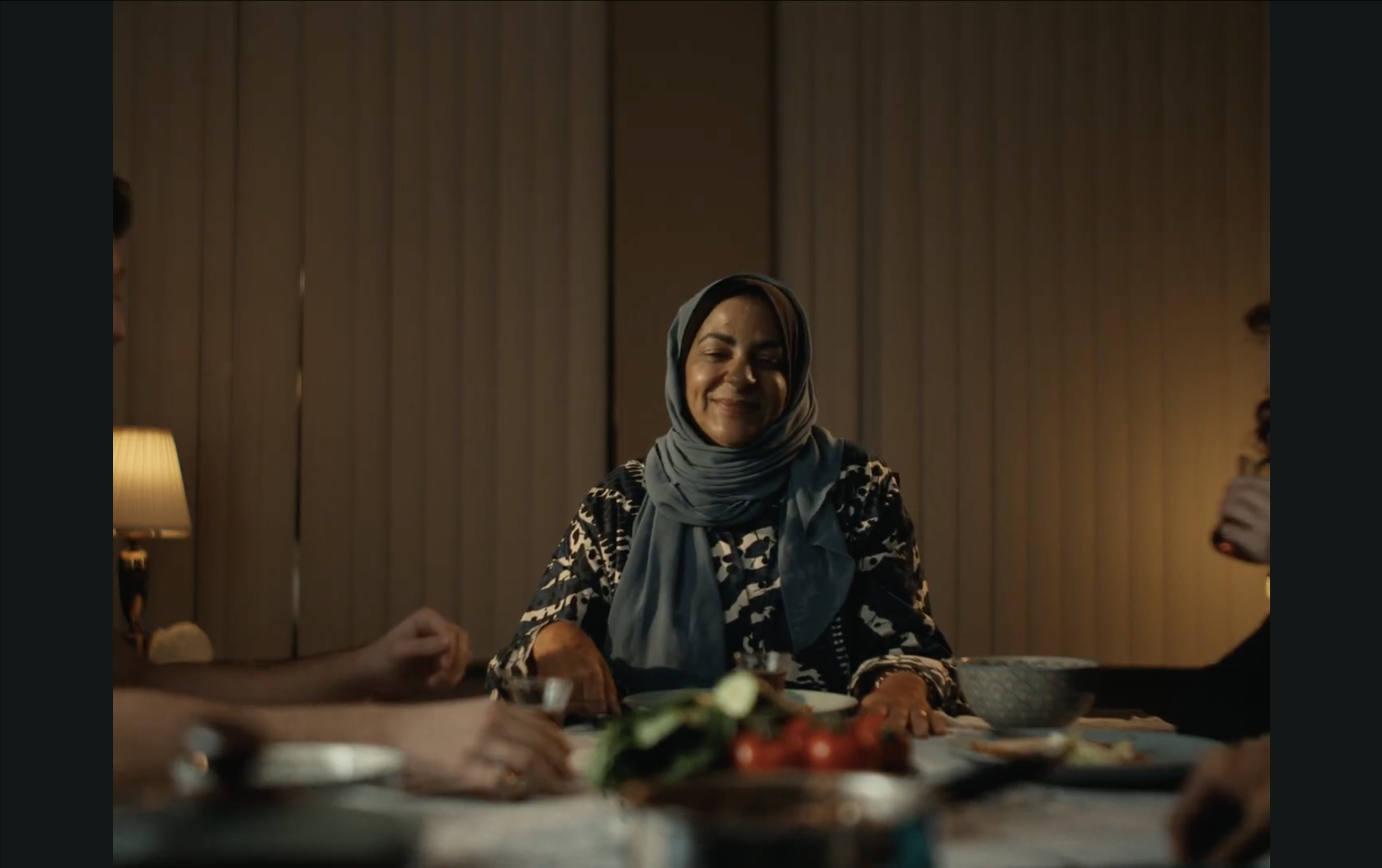 A woman wearing a headscarf and a patterned top smiling at a family dinner with food on the table.