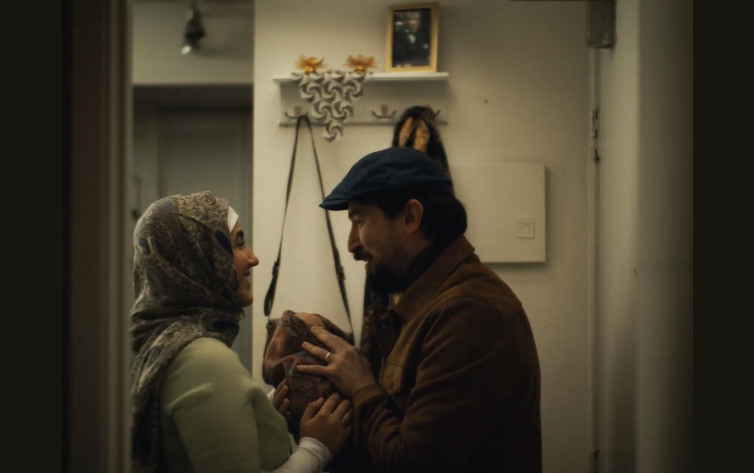 A man and a woman are standing close together in an intimate moment, holding hands. The woman is wearing a headscarf and the man is wearing a hat, and they are smiling at each other.