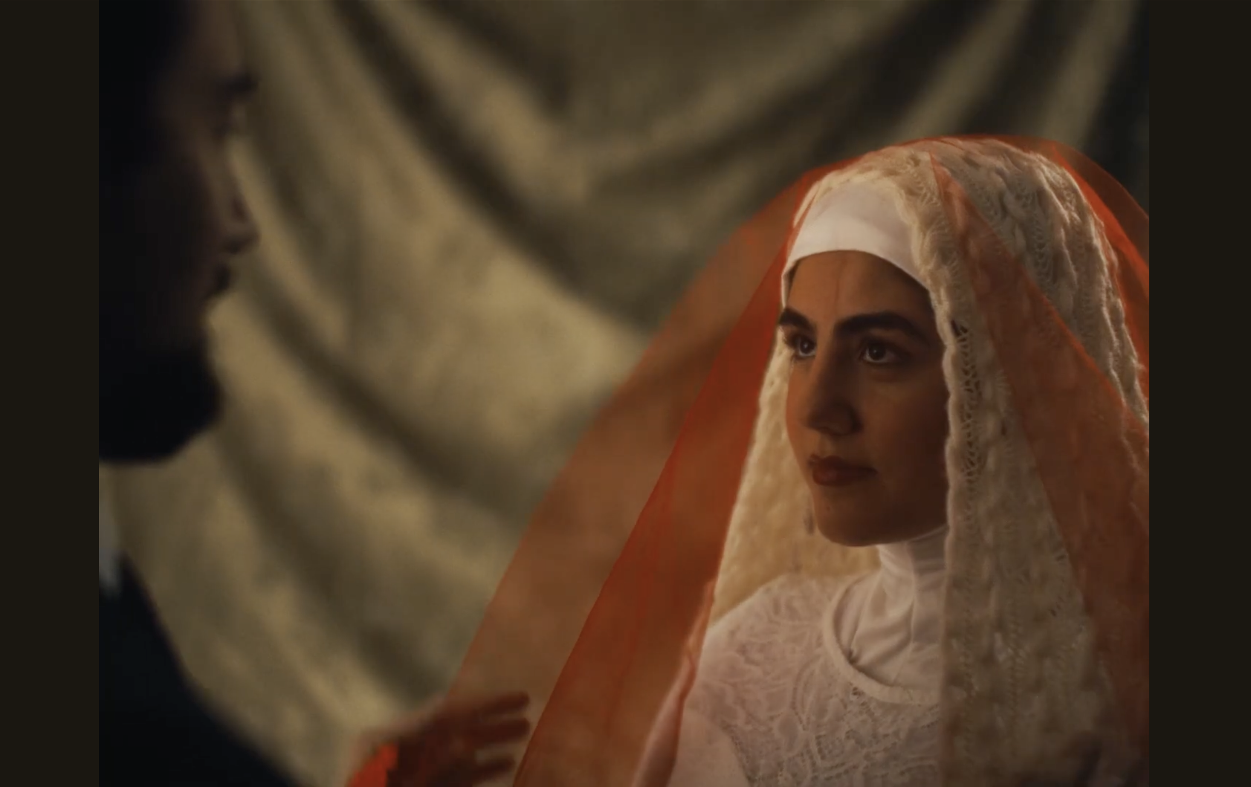 A woman dressed in traditional, elegant clothing with a head covering, looking intently at someone out of frame. The scene appears to be emotional or serious.