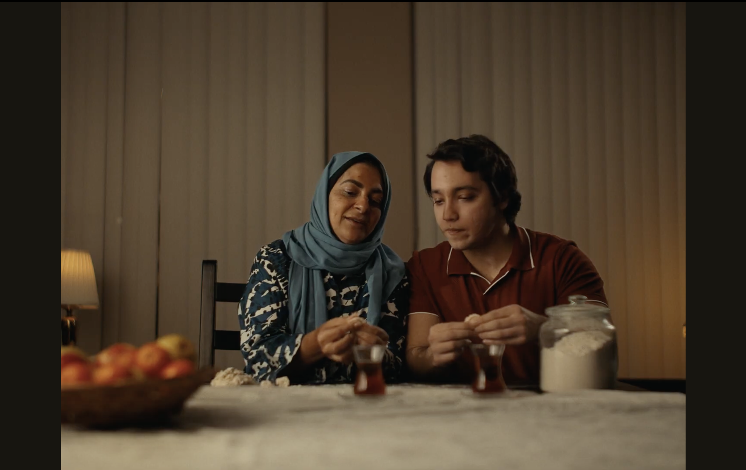 A woman wearing a blue headscarf and a young man with dark hair are sitting at a table, smiling and looking at something in their hands. The table has a glass jar of sugar and some dessert dishes. In the background, there are vertical wood panels and a small table lamp.