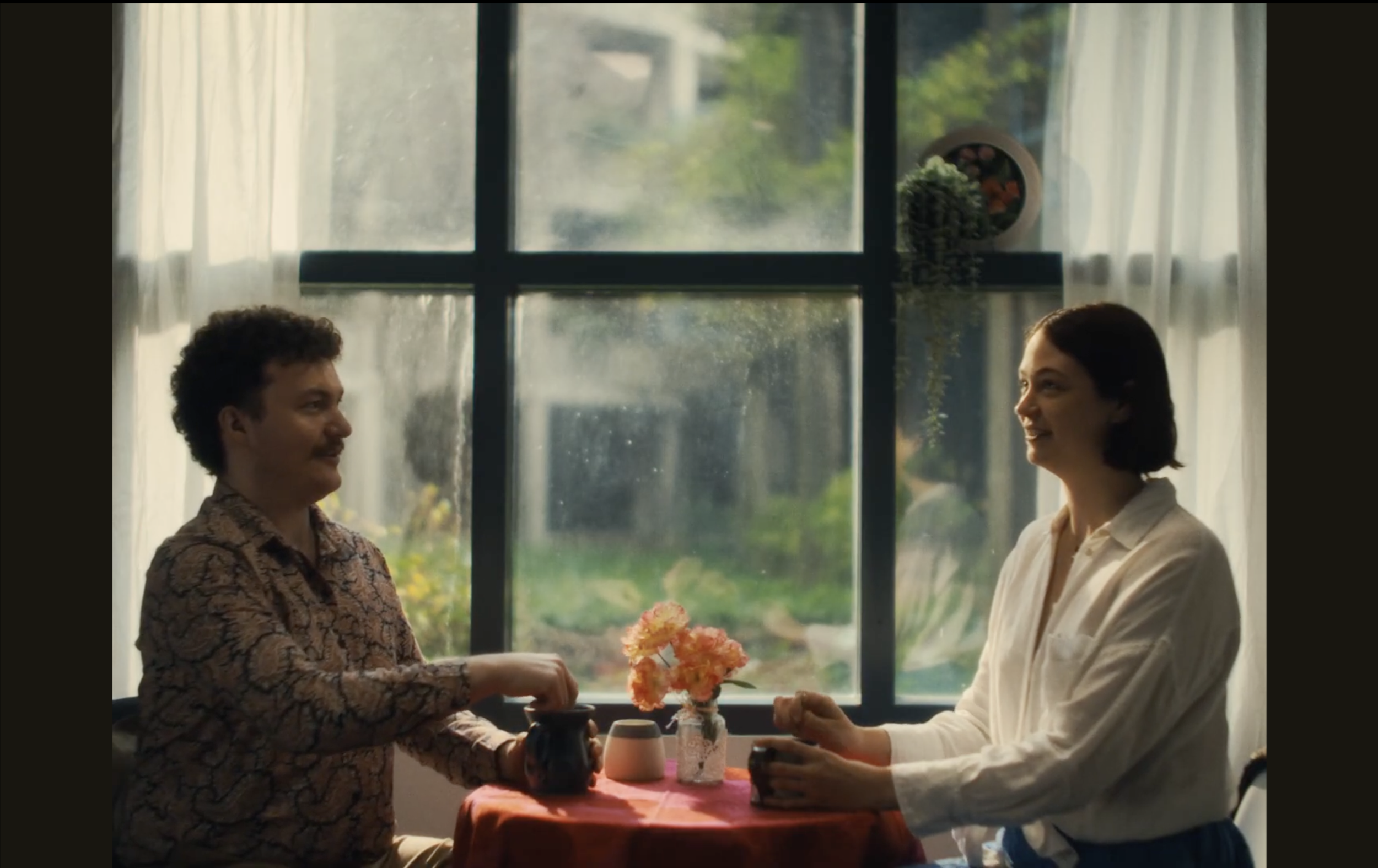 A man and woman are standing at a table, exchanging flowers in a room with large windows and sheer white curtains.