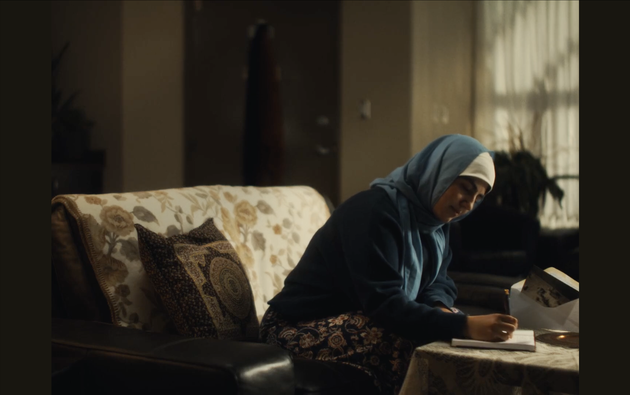 A woman wearing a white and blue headscarf sitting on a floral-patterned couch, writing a letter in a softly lit living room.