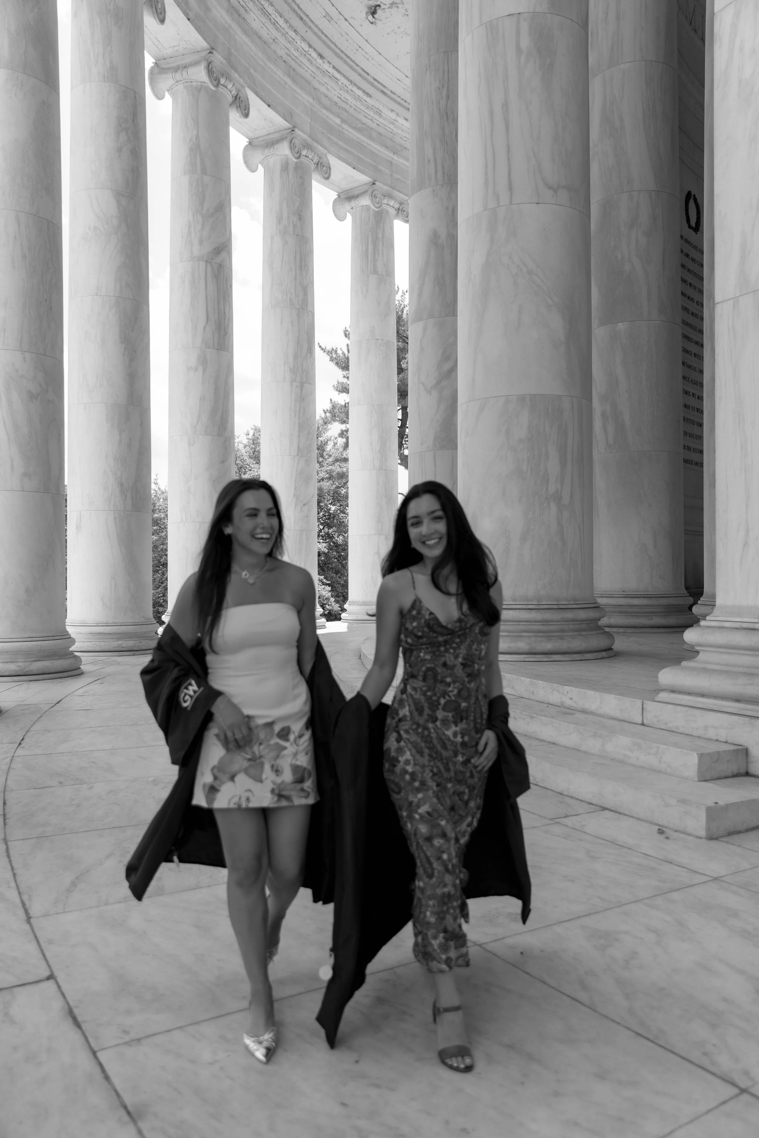 Two smiling women walking hand-in-hand in front of large marble columns at a historic building, dressed in formal attire with open academic gowns.