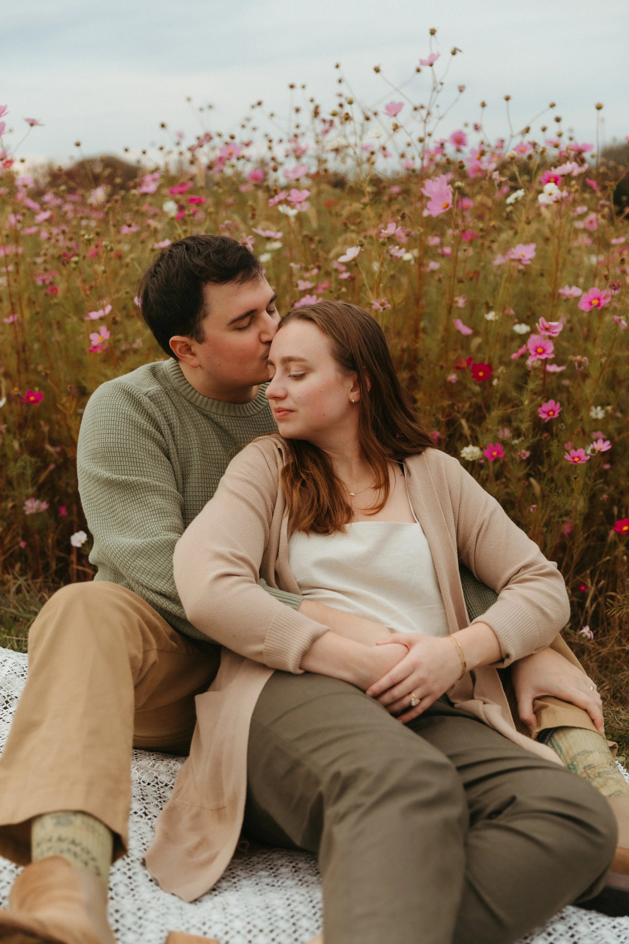 A couple sitting in a field of pink and white flowers, with the man kissing the woman's forehead.