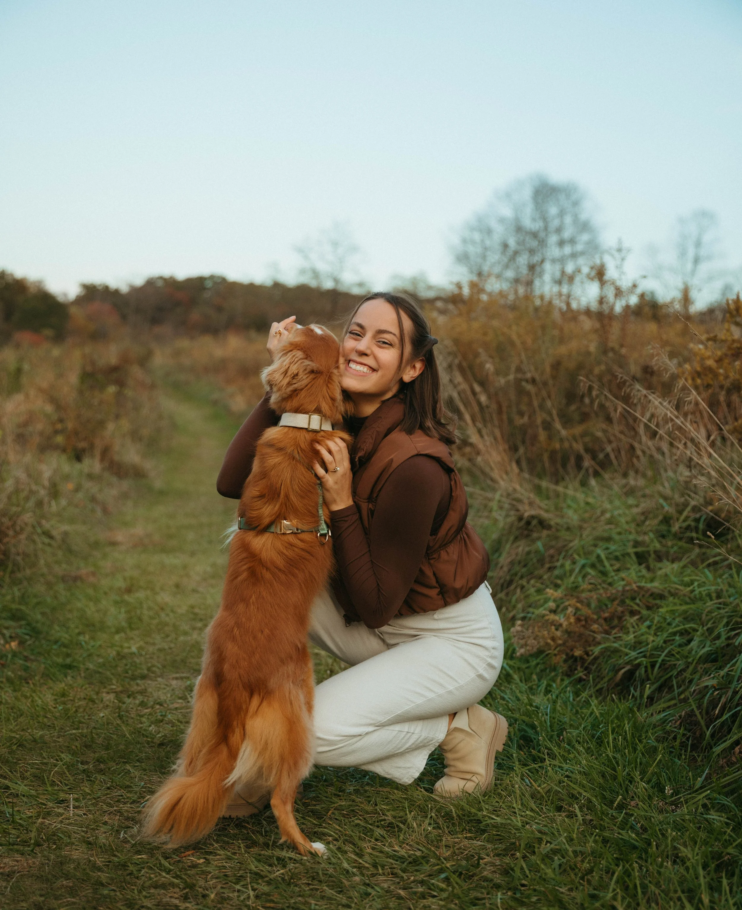 A woman hugging a golden retriever dog outdoors on a grassy trail surrounded by Autumn foliage.