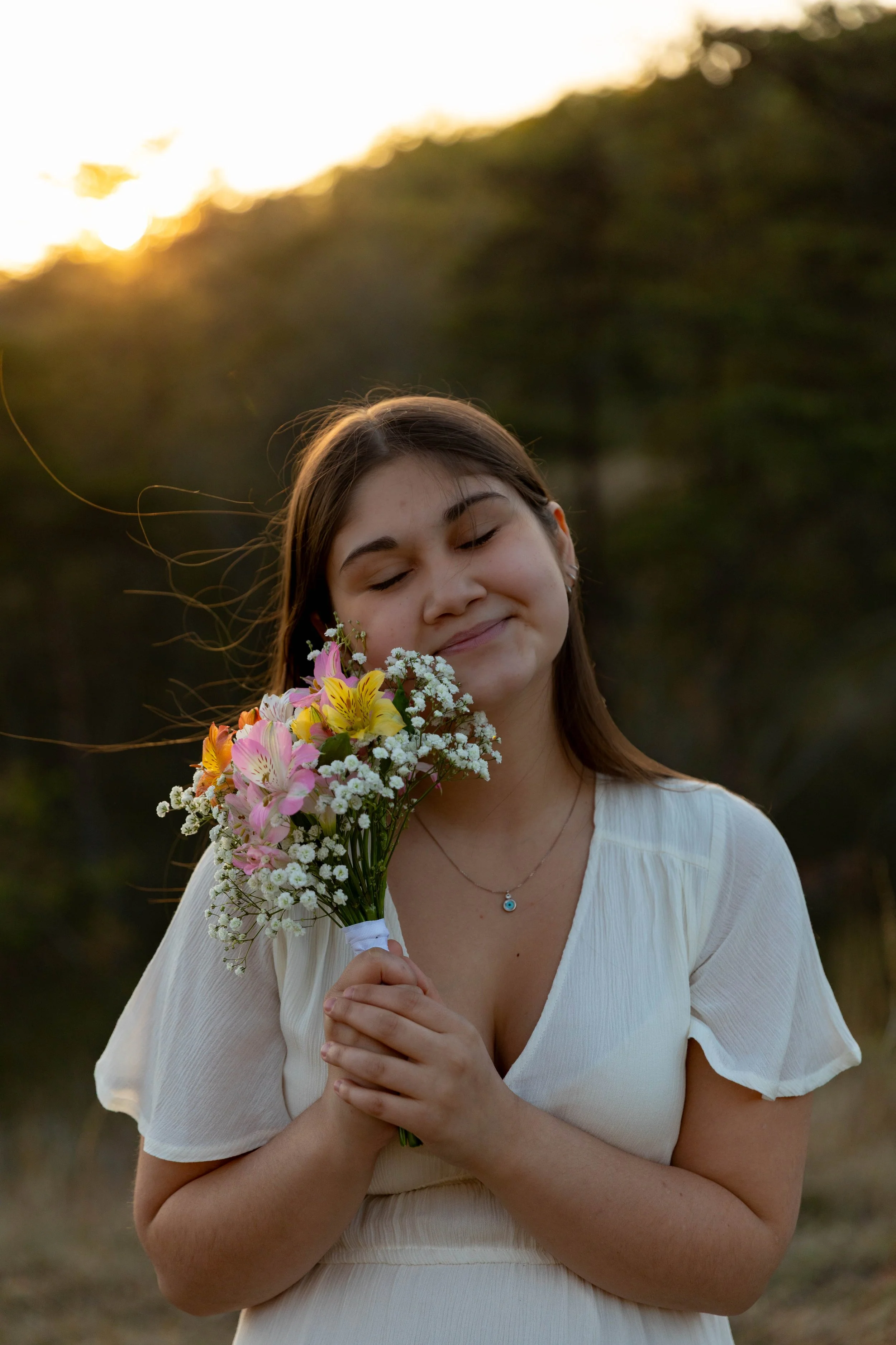 Young woman with long brown hair holding a bouquet of pink, yellow, and white flowers with green leaves, standing outdoors at sunset, with a soft smile, wearing a white dress and a silver necklace with a small pendant.