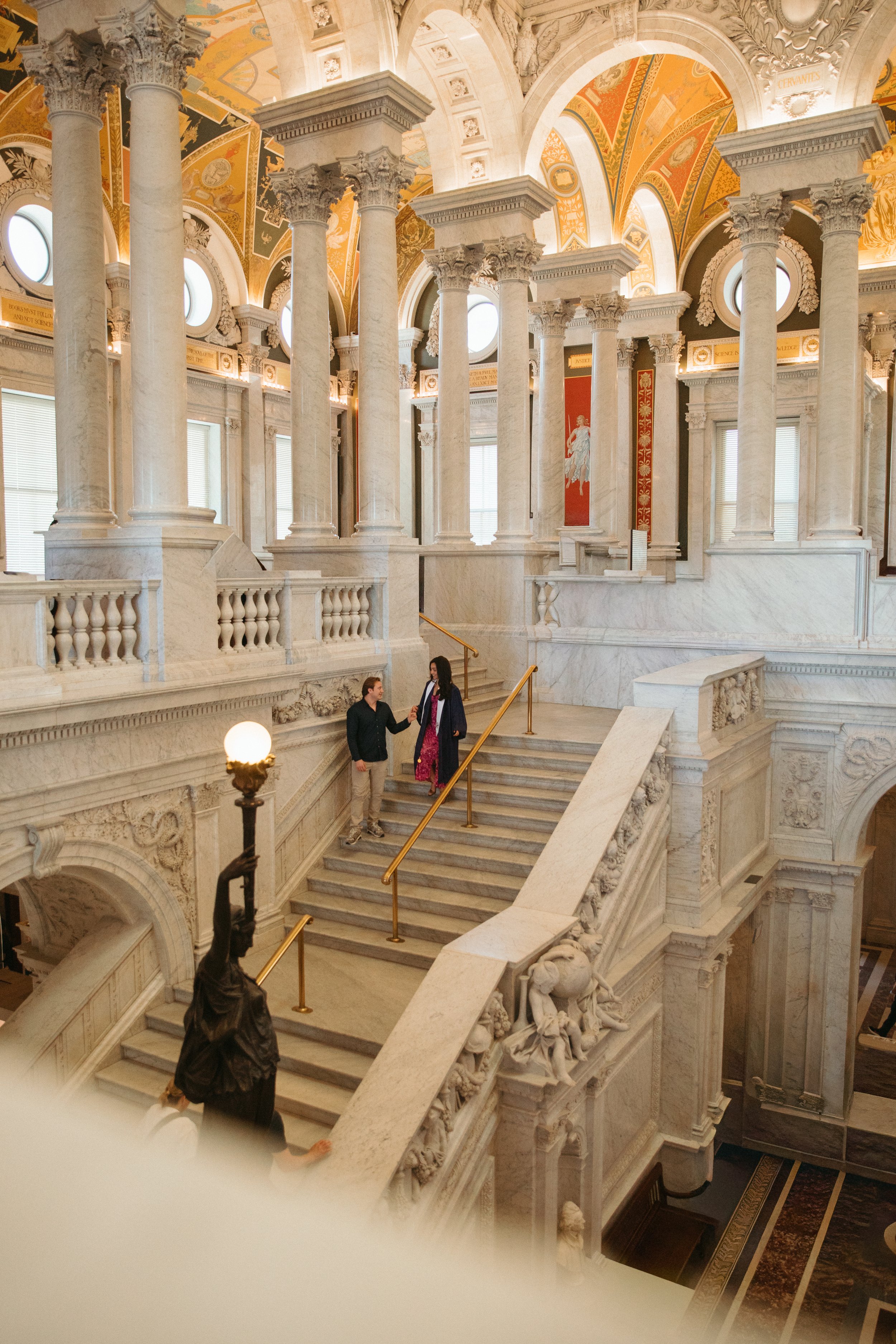 Interior of a grand, ornate building with marble stairs, large columns, decorative arches, and classical statues. Two people are conversing on the staircase.