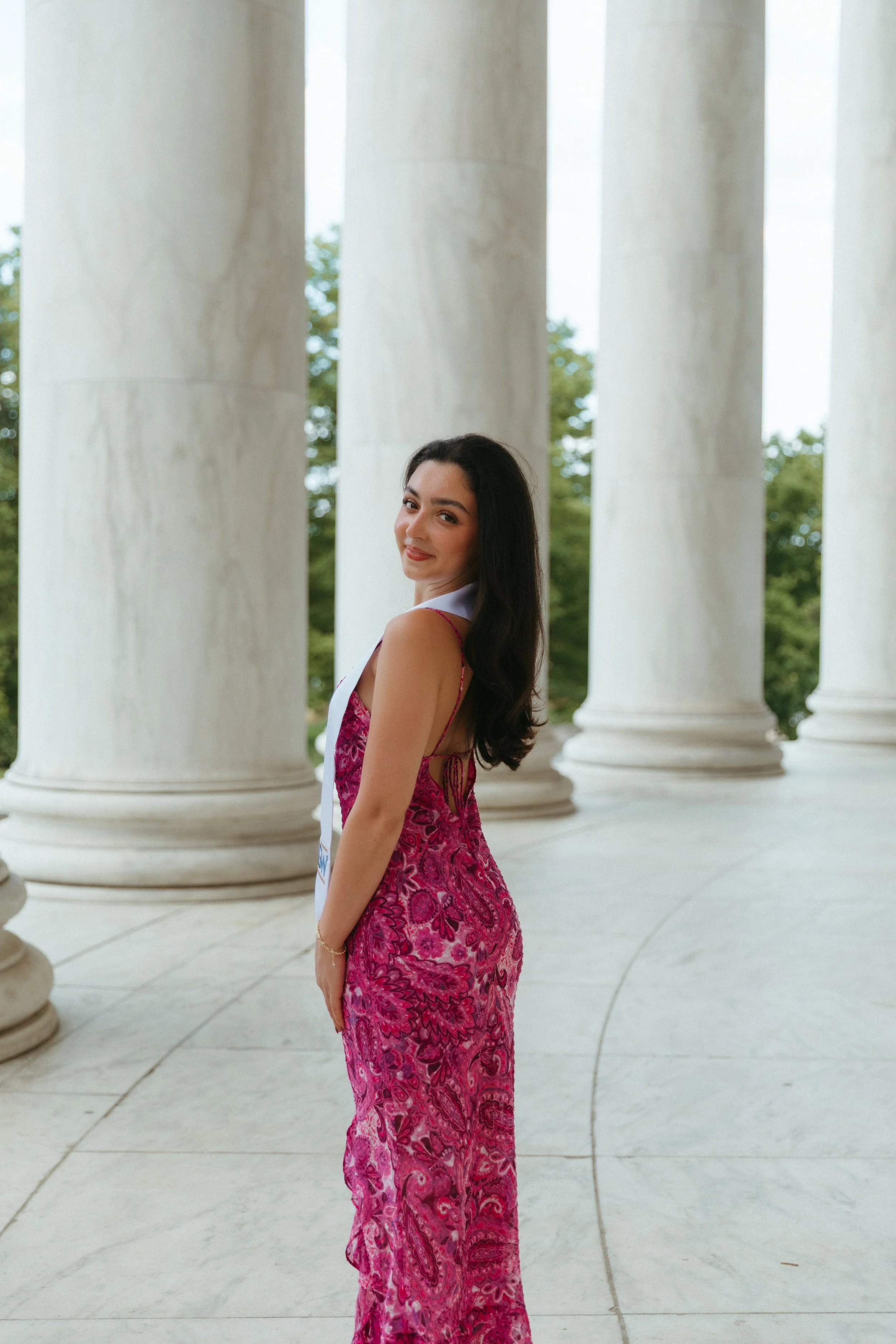 A woman in a pink patterned dress stands between large white columns outdoors, looking back and smiling.