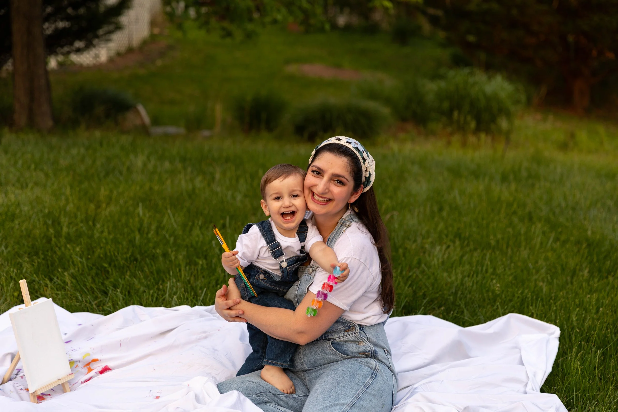 A young woman and a small boy sitting on a white cloth outdoors on a grassy area, smiling and hugging. The woman is wearing a white shirt, denim overalls, and a headscarf, while the boy is dressed in a white shirt and denim overalls, holding paintbrushes.