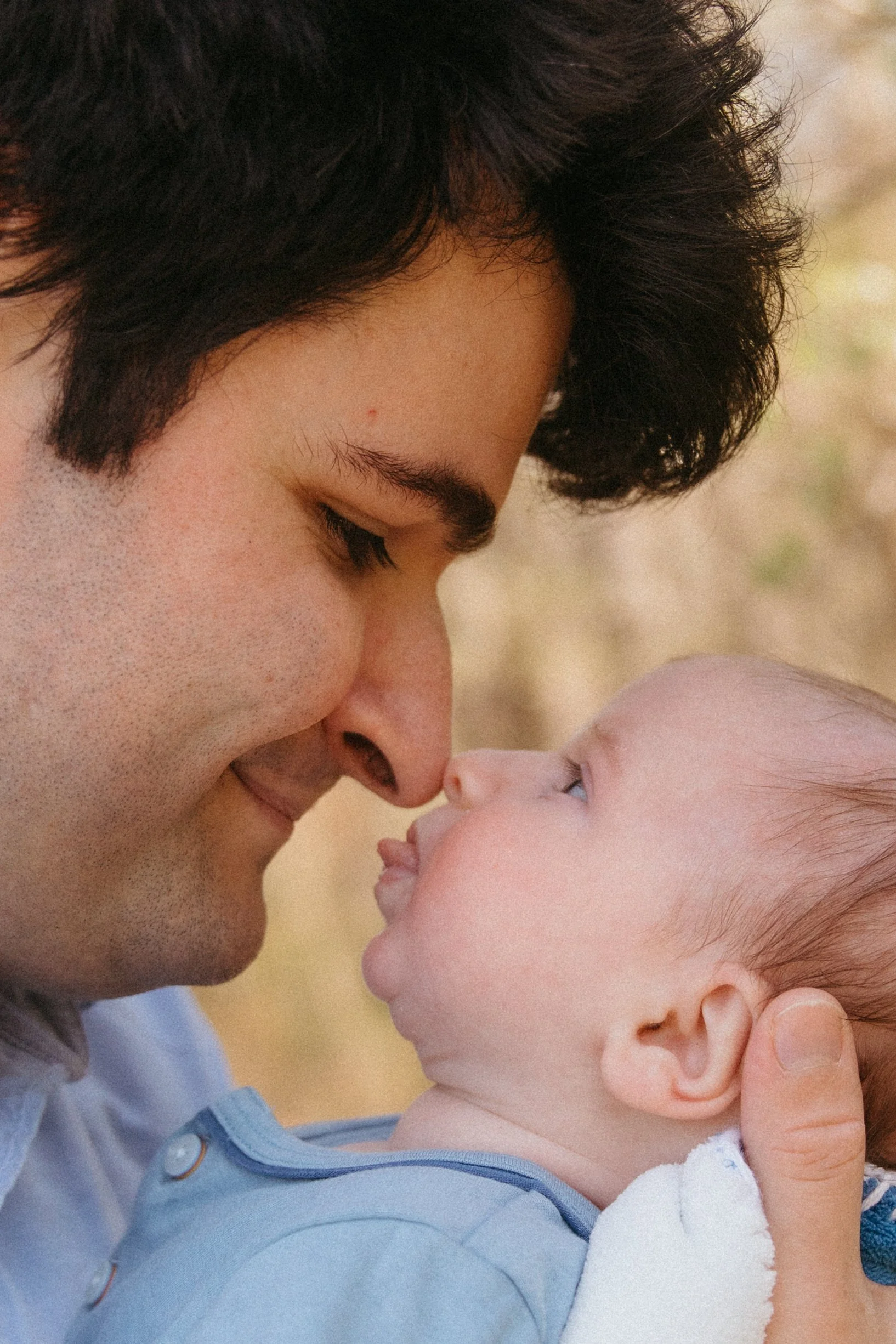 A close-up of a man and a baby, touching their noses together affectionately, outdoors with a blurred natural background.