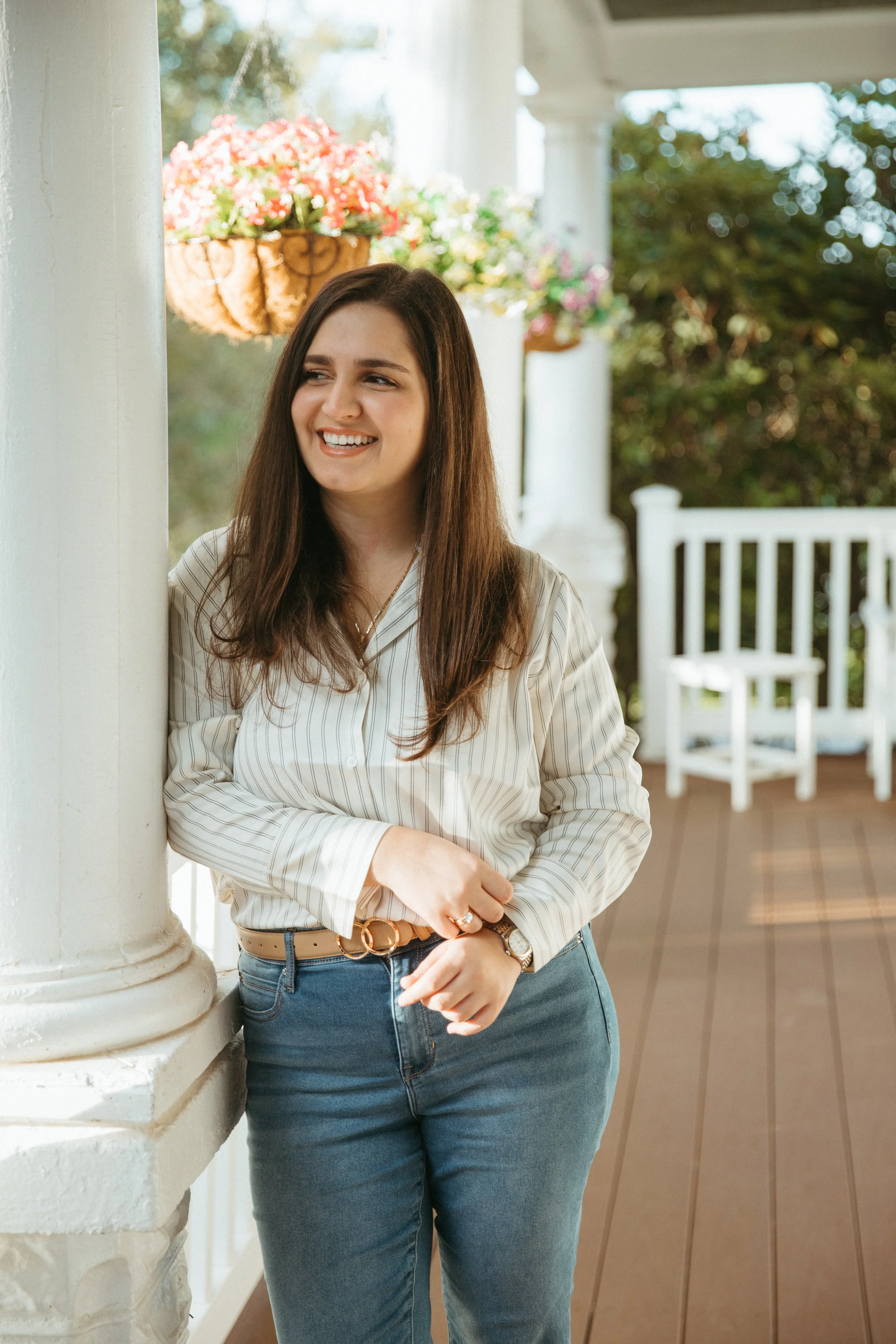 A woman with long brown hair, wearing a striped white and beige blouse and blue jeans, stands on a porch with white pillars, smiling and looking to the side.