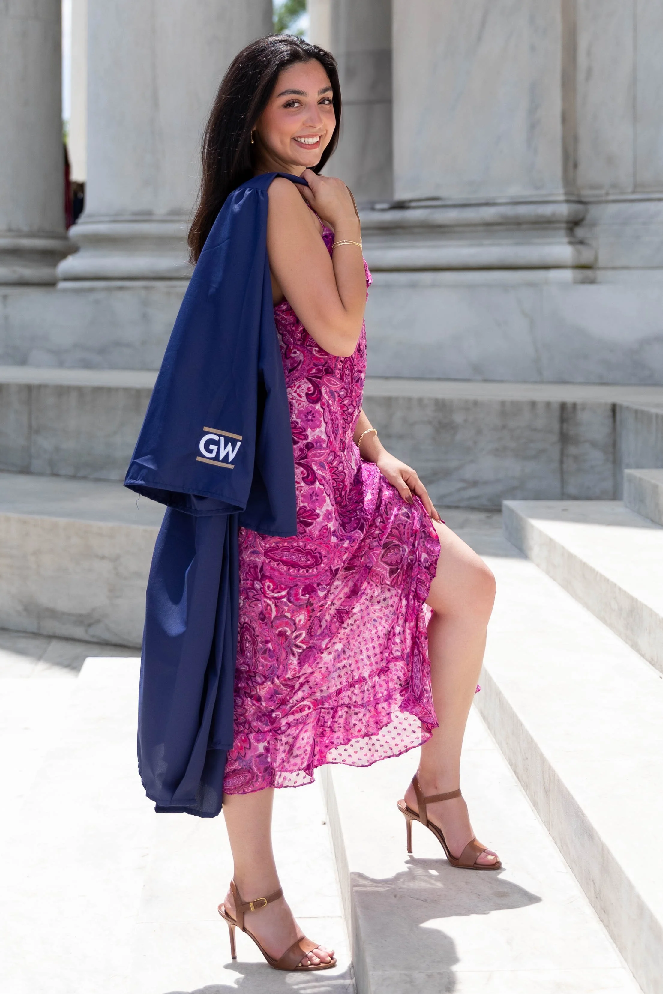 A woman in a pink dress sitting on marble steps, smiling, with a navy blue jacket draped over her shoulder.