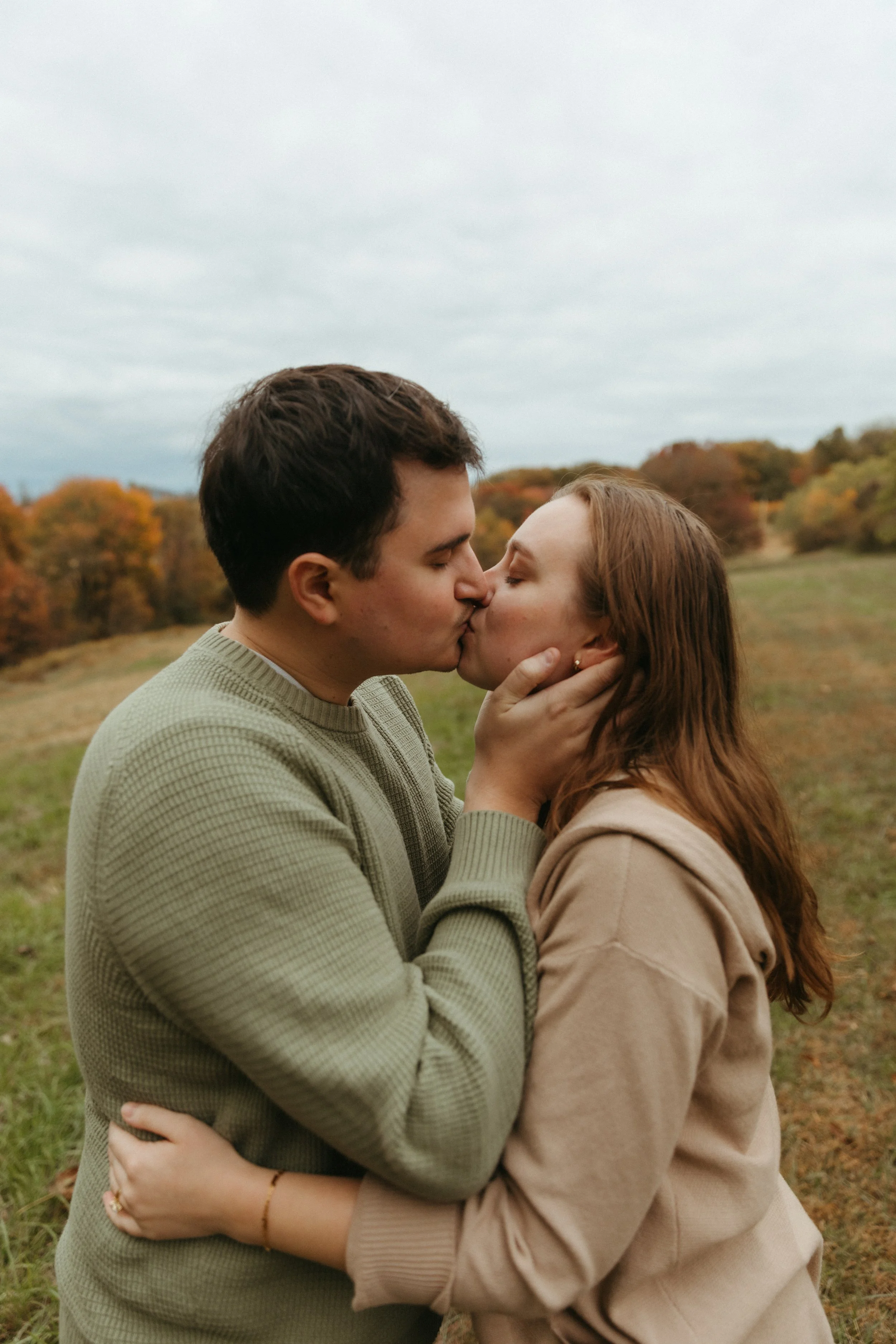 A couple sharing a kiss outdoors in a grassy field with trees showing autumn colors.