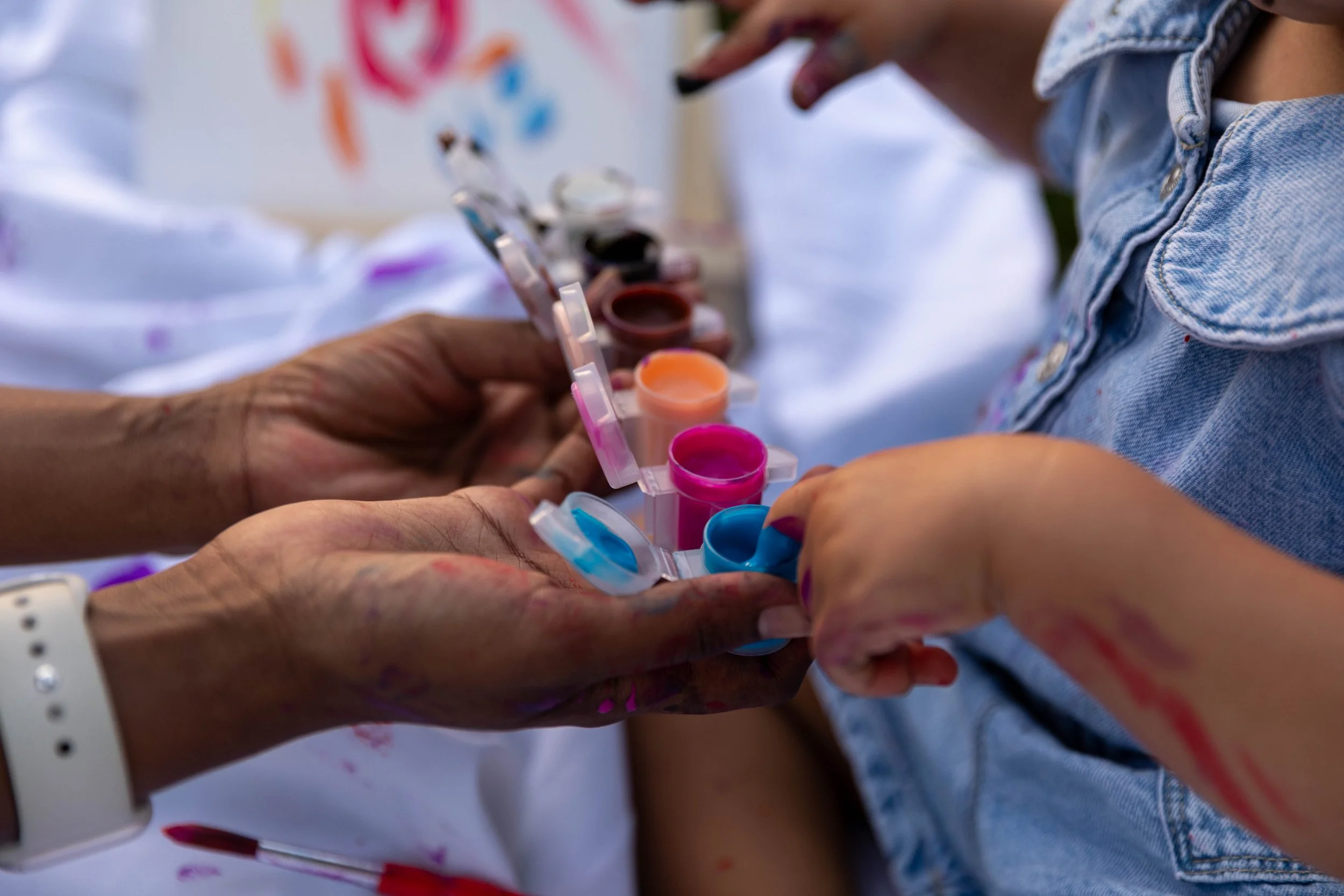 Close-up of a child's arm being painted with colorful face or body paints by an adult using a small paint palette with various colors.