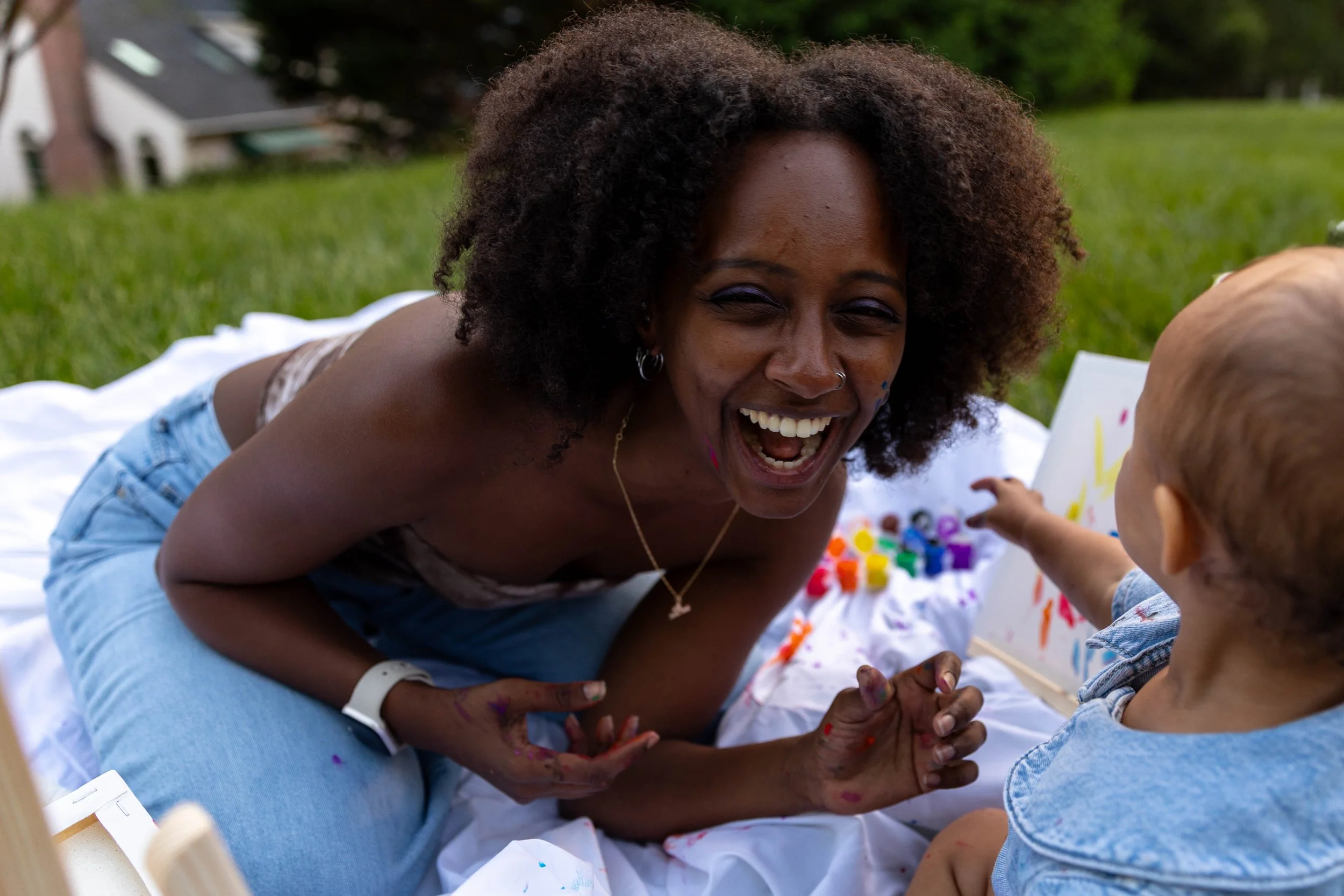 A woman with curly hair and a big smile, laughing and playing with a young child outdoors on a grassy area during the daytime.
