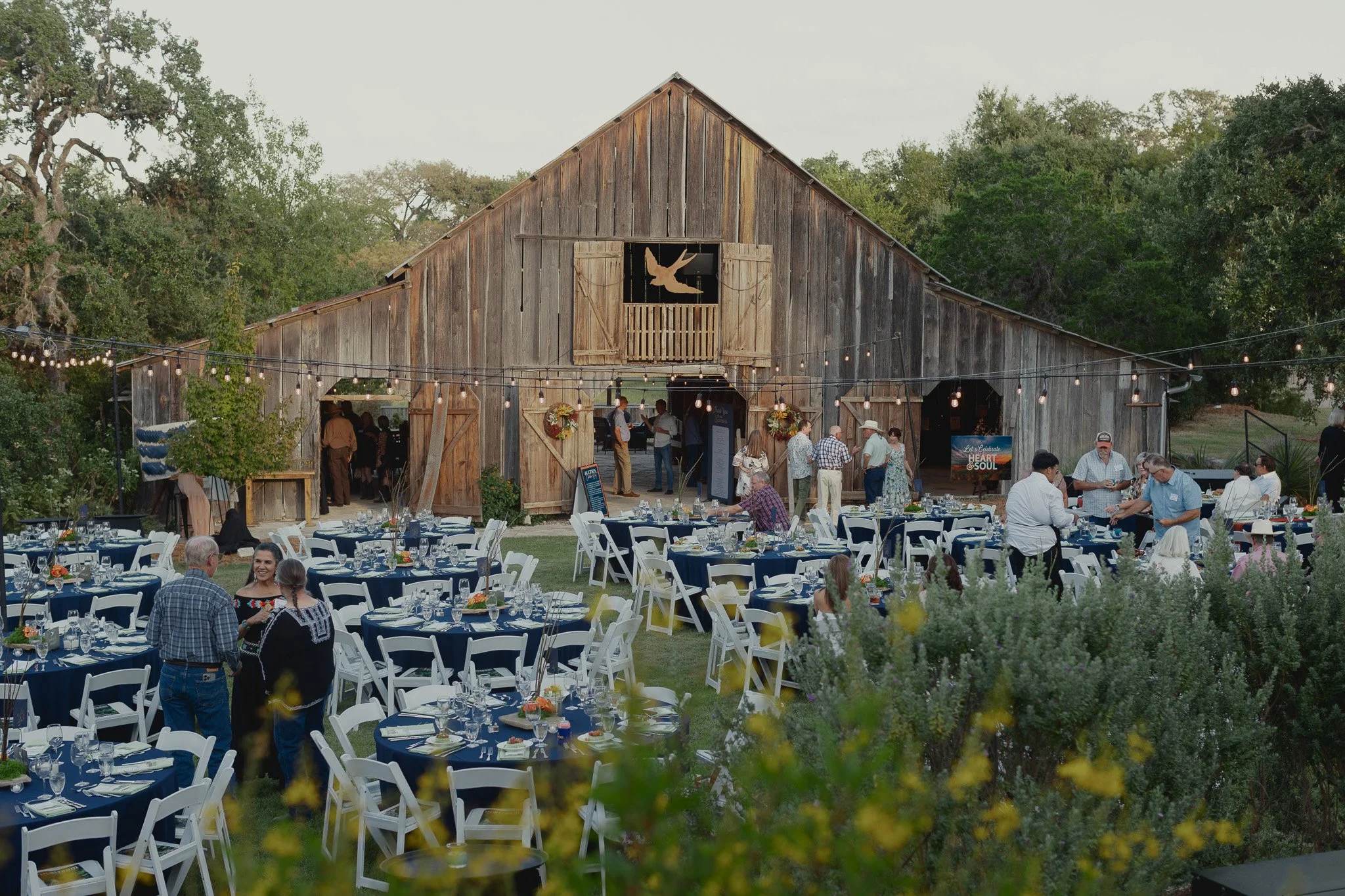 Outdoor event at a rustic barn with tables set for a celebration, surrounded by greenery and string lights.