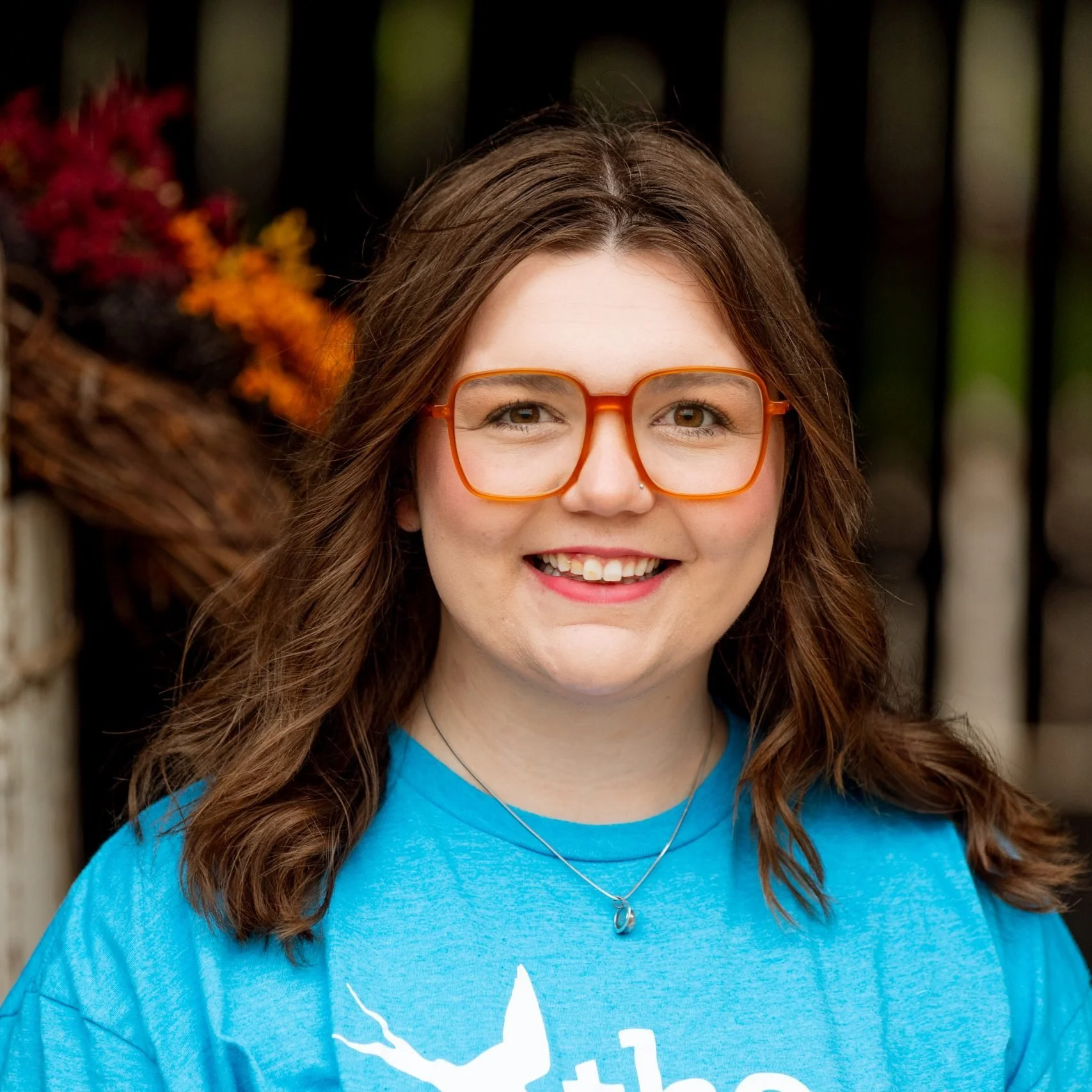 A young woman with wavy brown hair, smiling, wearing orange glasses, a blue t-shirt, and a necklace, standing outdoors in front of a wooden fence with colorful flowers on the left.