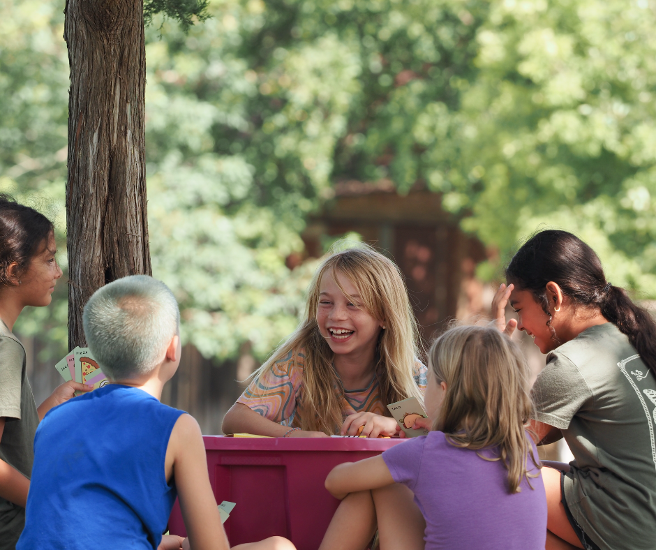 A group of children playing a card game outdoors in a park with green trees and sunlight.