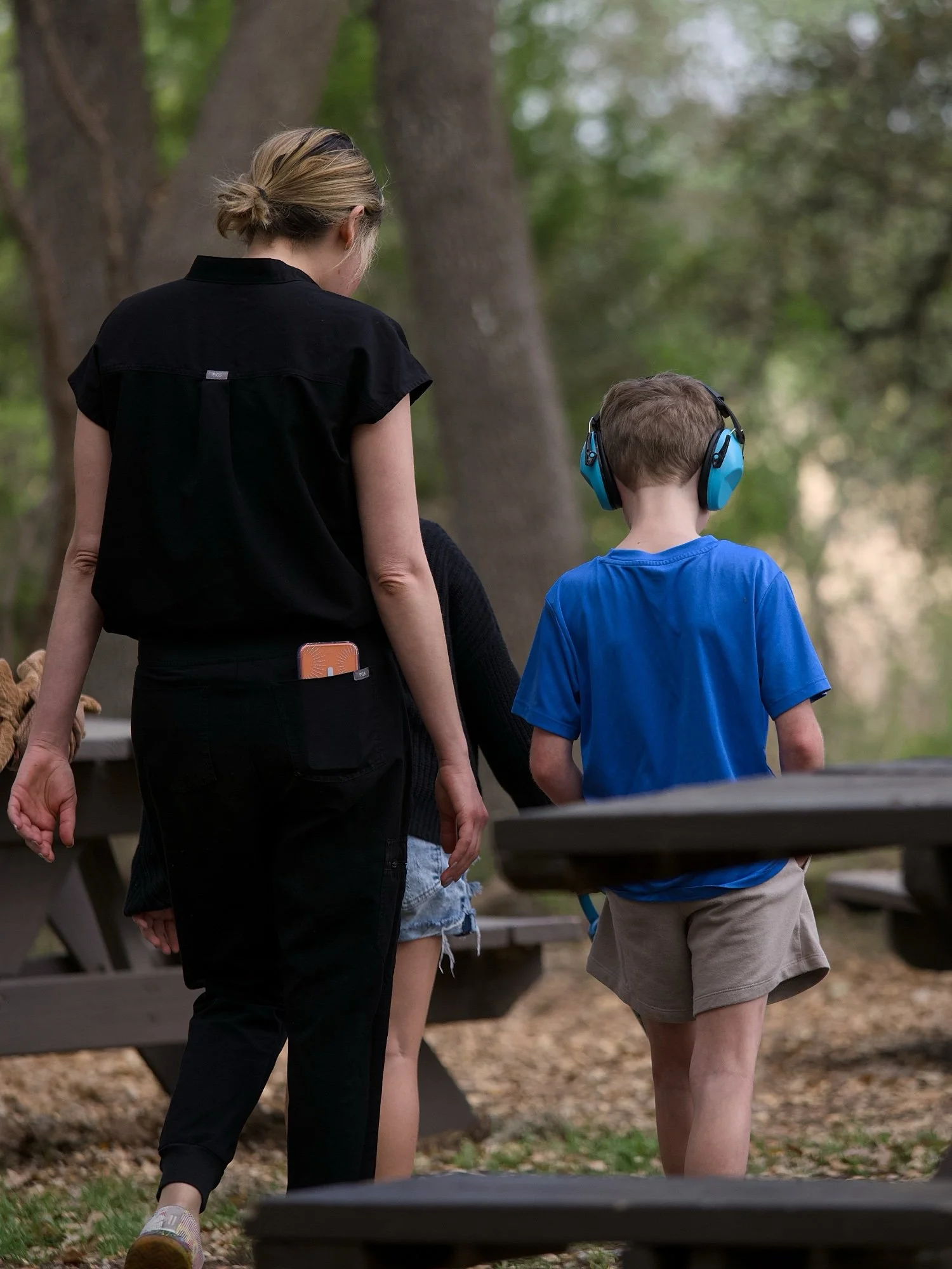 A woman and a young boy walking in a park with tall trees and fallen leaves, with the boy wearing blue headphones and a blue shirt.