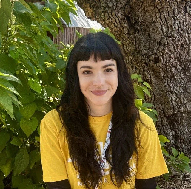 Smiling woman with brown hair wearing a blue t-shirt, standing outside near a wooden fence.