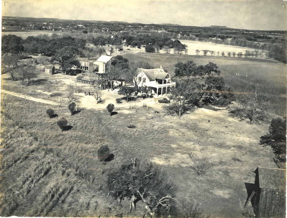 A rural landscape featuring a large farmhouse with a porch, several trees, a dirt road, and a barn or shed nearby, with fields stretching into the distance.