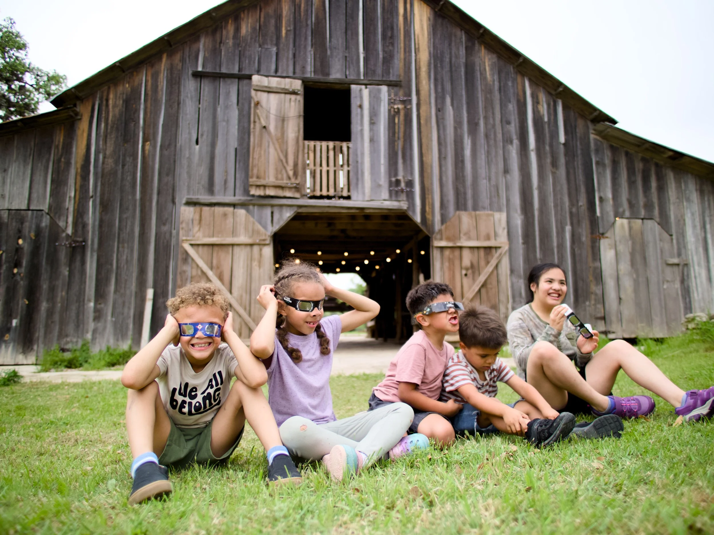 A group of children sitting on grass in front of a wooden barn, some wearing solar eclipse glasses, smiling and enjoying outdoors.