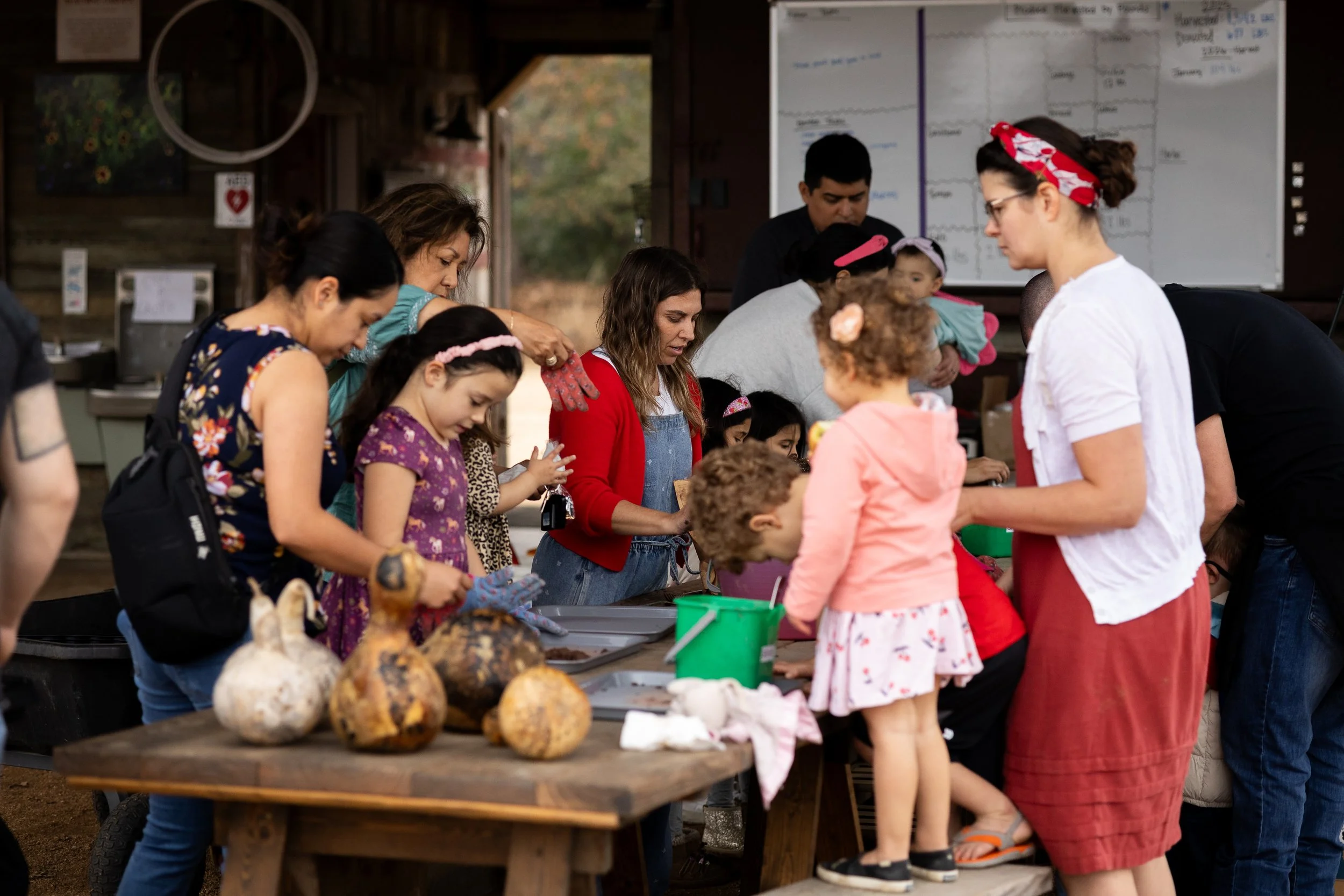 A group of children and adults gathered around a table inside a rustic barn, with vegetables and art supplies on the table, engaged in a craft activity.