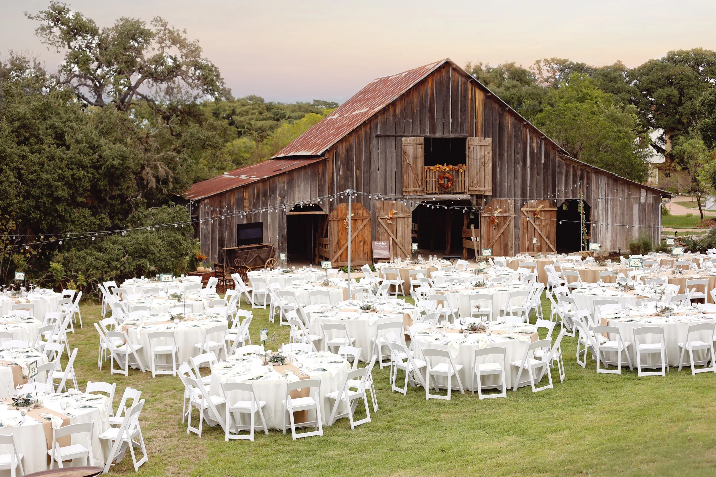 Rustic barn with tables and chairs set up for an outdoor event, surrounded by trees and greenery with string lights hanging across the area.