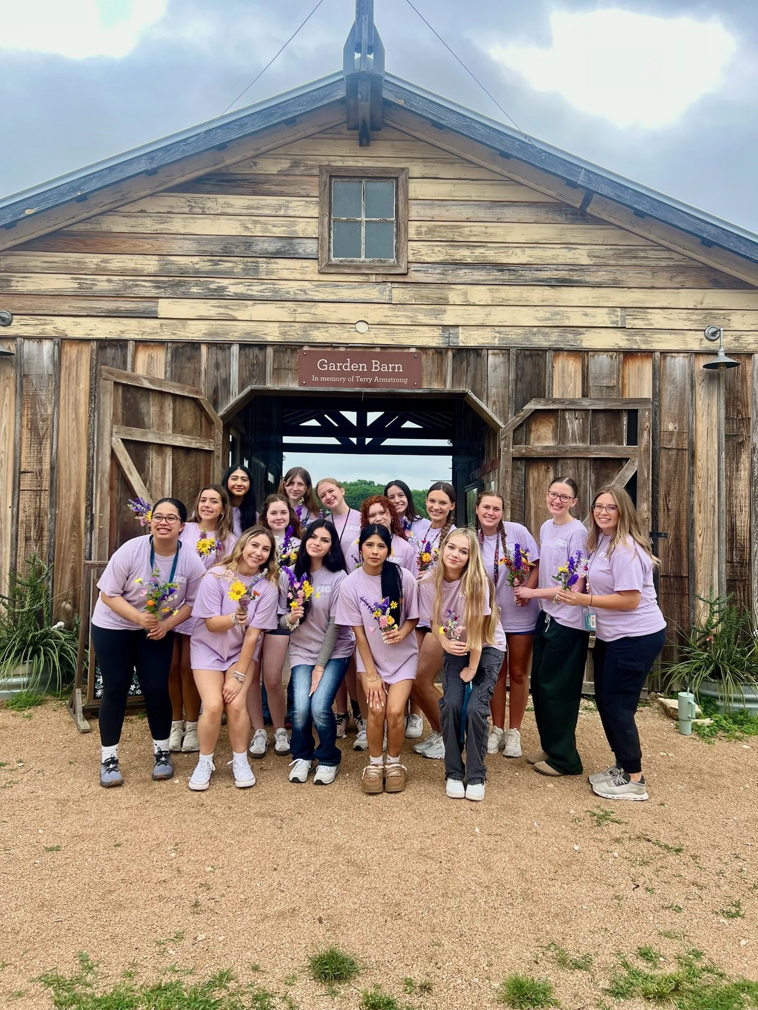 A group of young women standing in front of a rustic wooden barn, holding bouquets of flowers, smiling, and wearing matching lavender T-shirts.