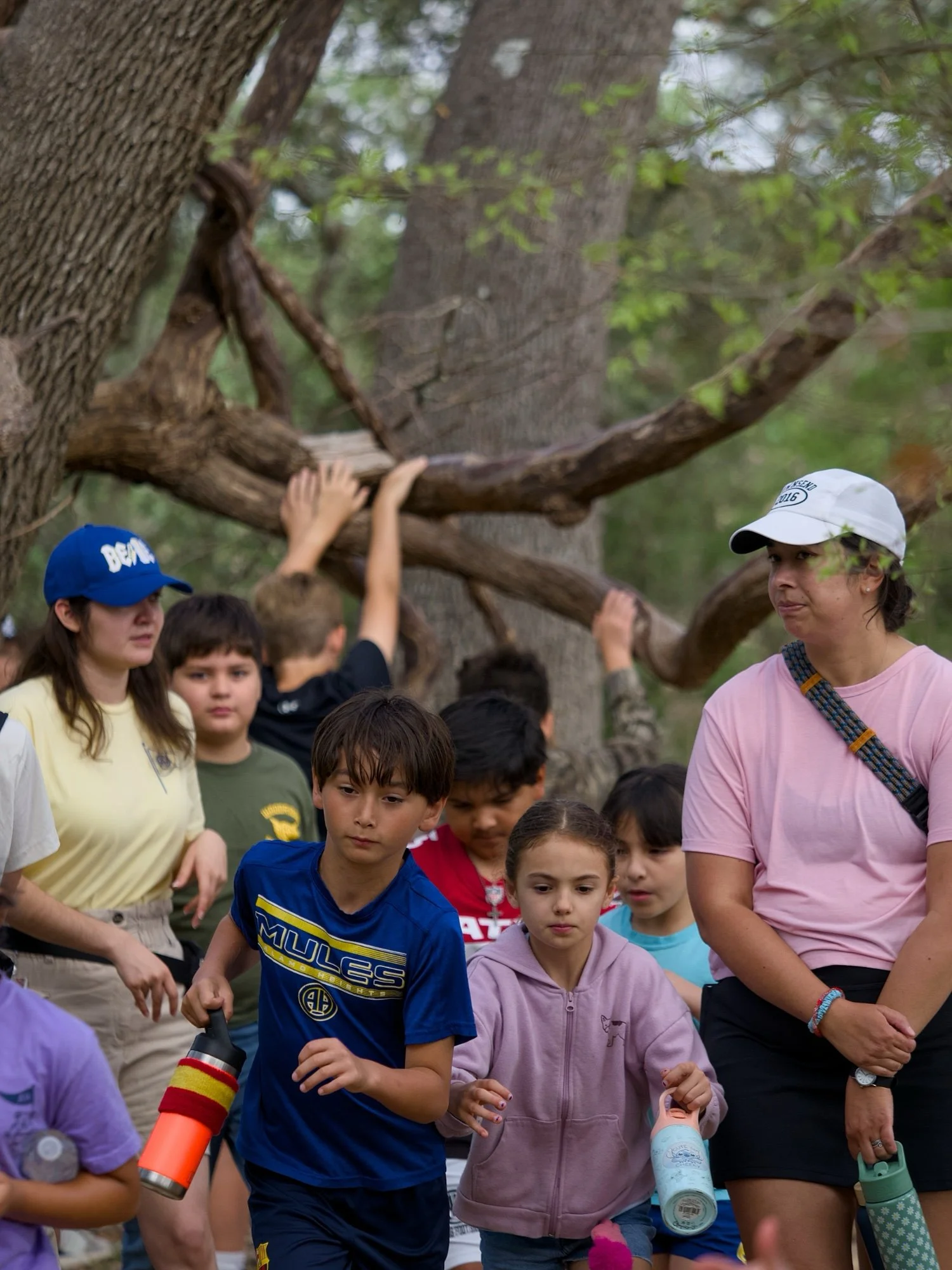 Group of children and women outdoors, some holding water bottles, near a large tree with green leaves.