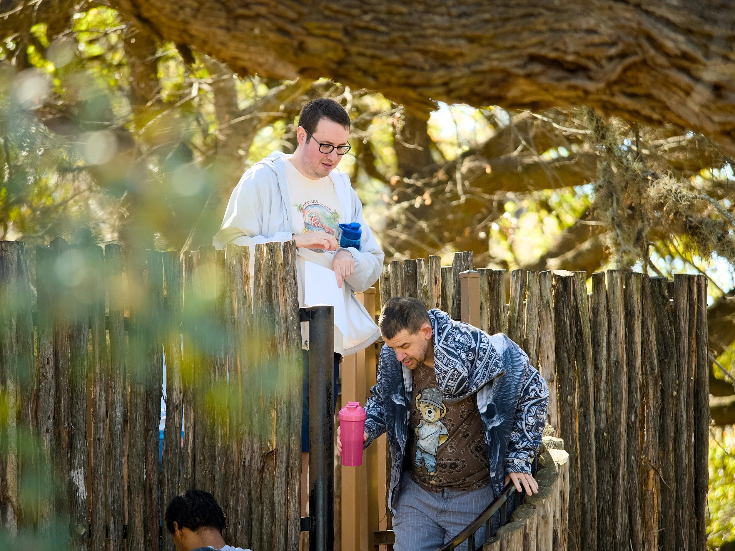 Group of people by a wooden fence in a park with trees, a young man with glasses wearing a white hoodie standing behind the fence, and others wearing patterned jackets, one holding a pink water bottle.
