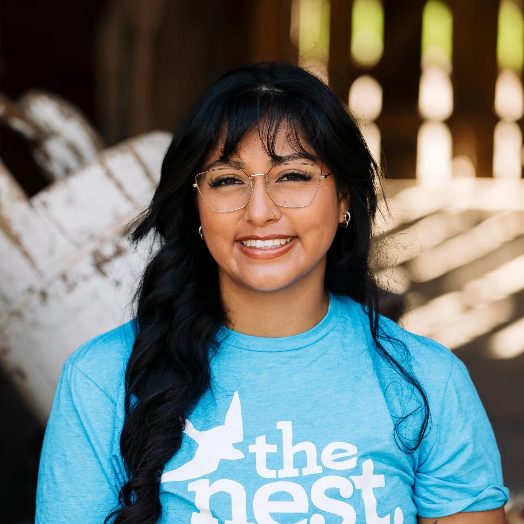 A young woman with black hair, glasses, and a nose ring smiling outdoors. She is wearing a blue t-shirt with white text and a white star graphic, and the background shows a rustic wooden fence with sunlight filtering through.