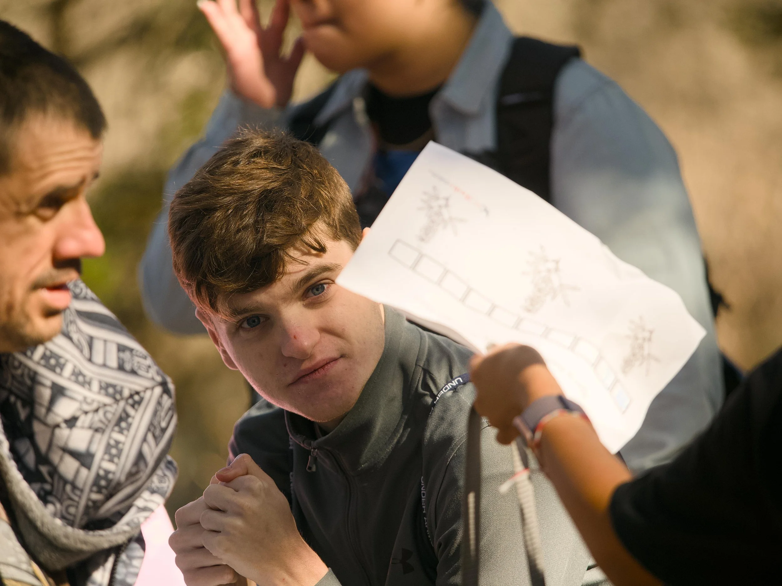 Two young men are engaged in a friendly arm-wrestling match outdoors, with one looking determined and focused, and the other observing. Two adults are also present, one on each side, watching the competition.