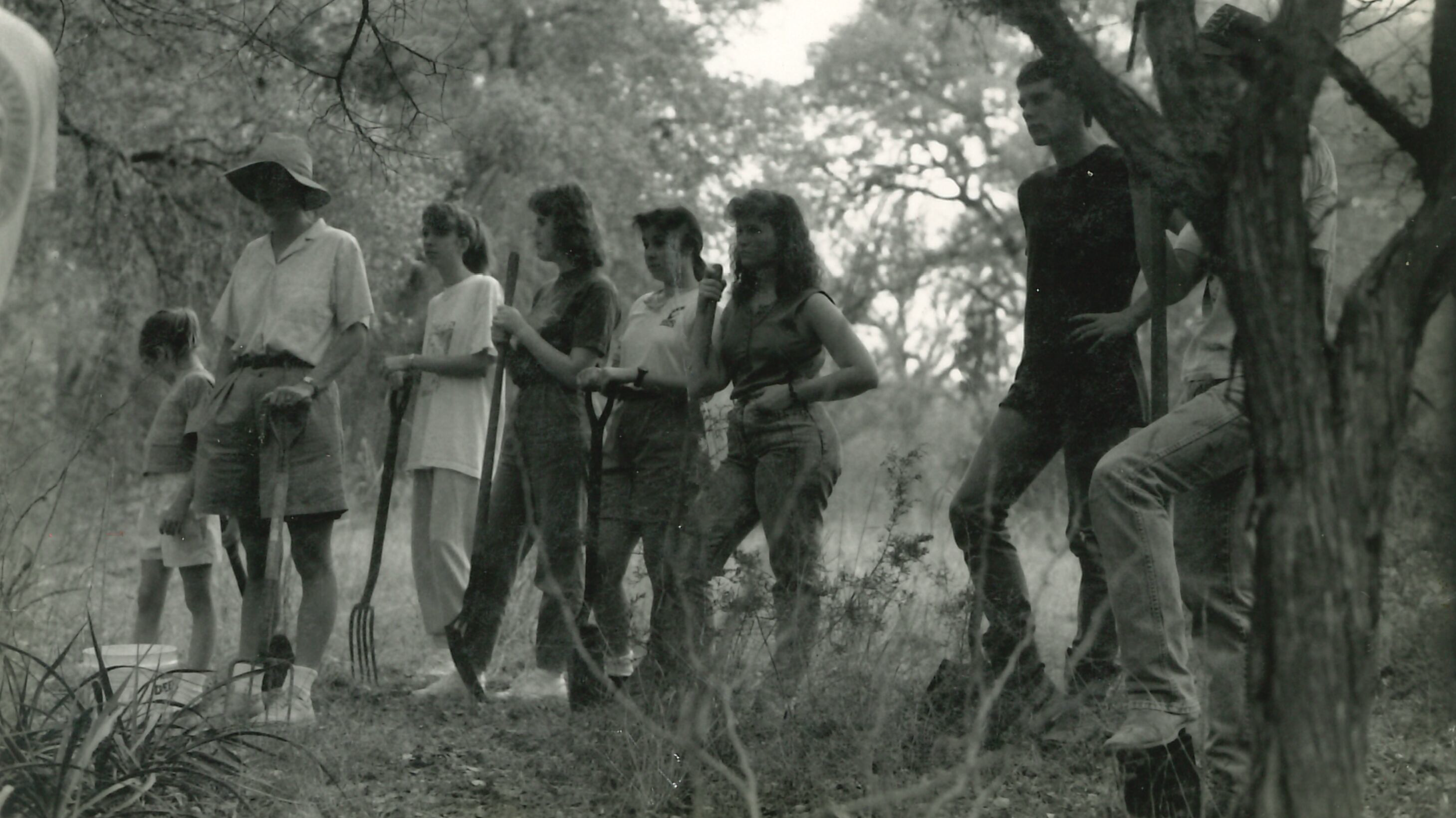 Group of people standing outdoors among trees, some holding tools, with a boy and girl in the foreground, black and white photograph.
