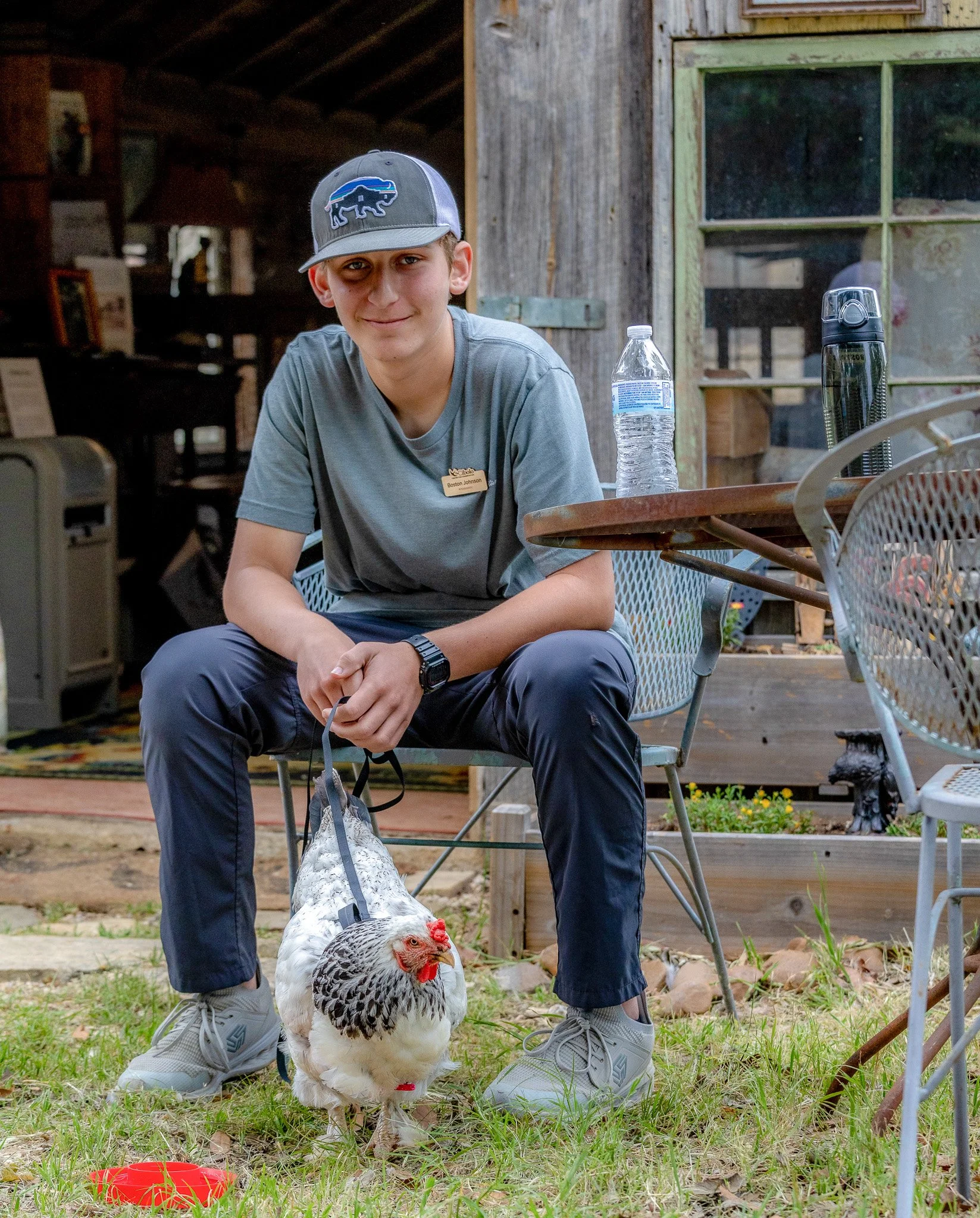 A young man wearing a gray cap and gray shirt, sitting outdoors, with a chicken standing in front of him. The background shows a rustic wooden building and some outdoor chairs. The man has a name tag that reads 'Boston Johnson'.