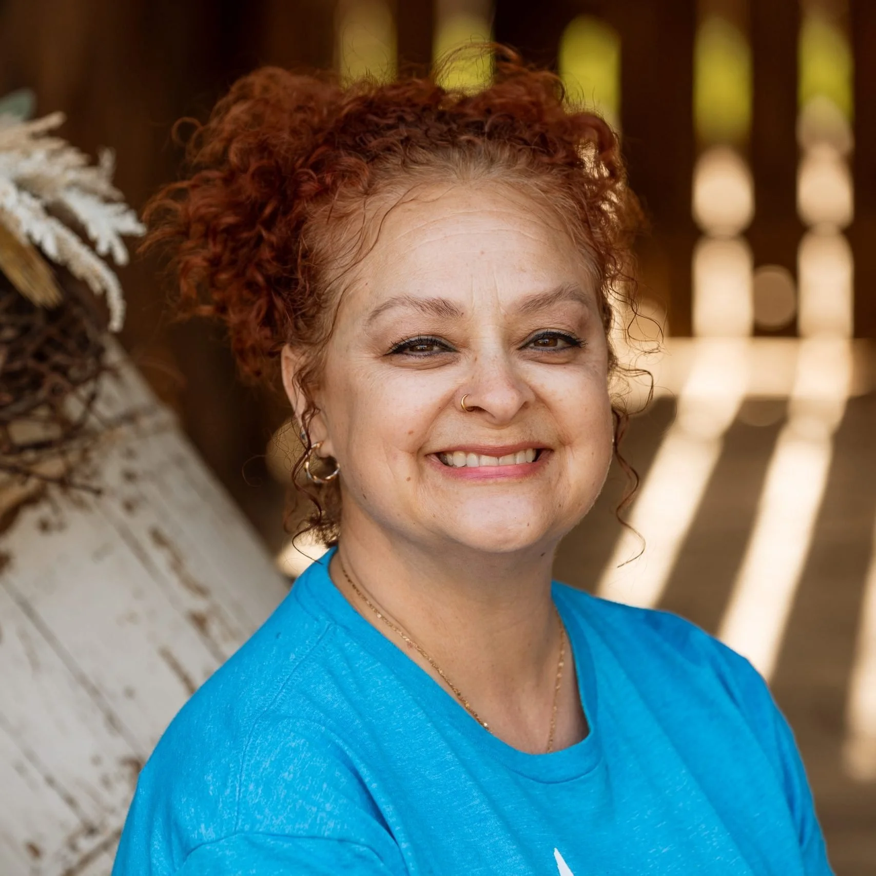 Close-up of a woman with curly red hair, wearing a blue shirt, smiling outdoors near a wooden fence with sunlight filtering through.