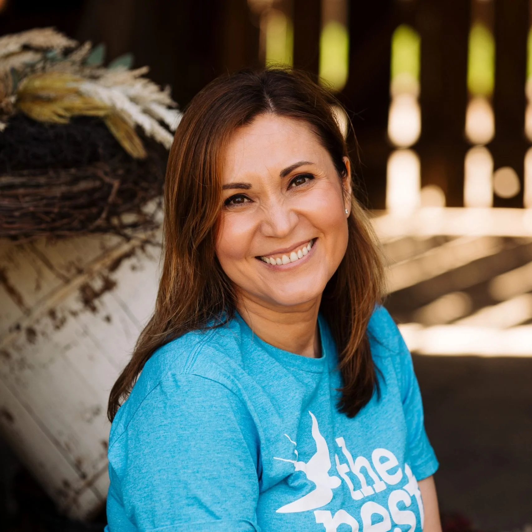 Smiling woman with brown hair wearing a blue t-shirt, standing outside near a wooden fence.