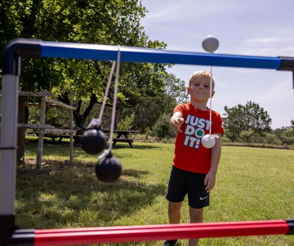 A young boy in a red t-shirt and black shorts playing a game of badminton outdoors. He is holding a white shuttlecock, and there are two black shuttlecocks hanging from a badminton net. The background includes trees, a picnic table, and a grassy fiel