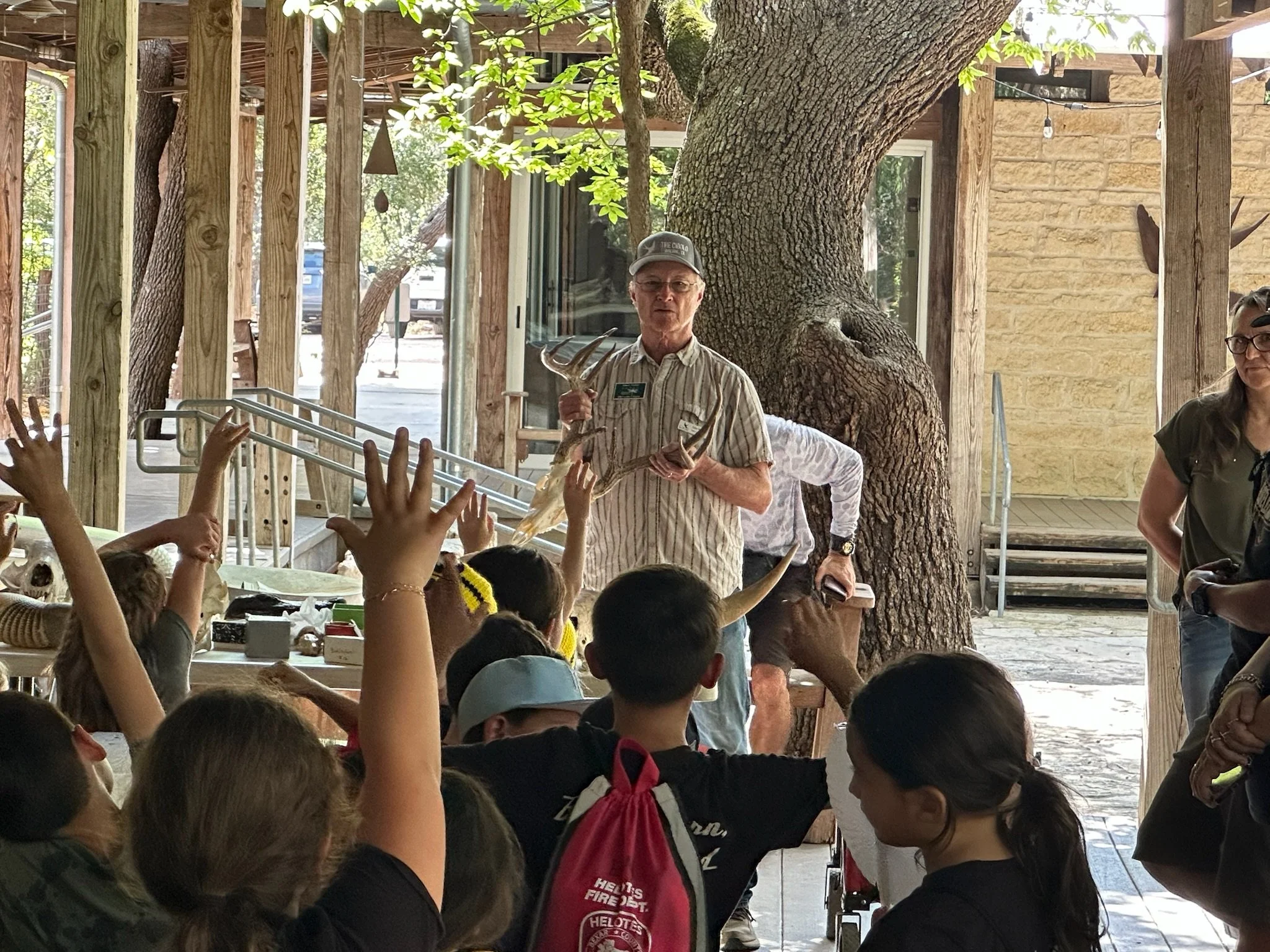 A man with glasses and a cap holding antlers, standing among children with raised hands, in an outdoor setting with large trees and wooden structures, during a field trip at the nature center.