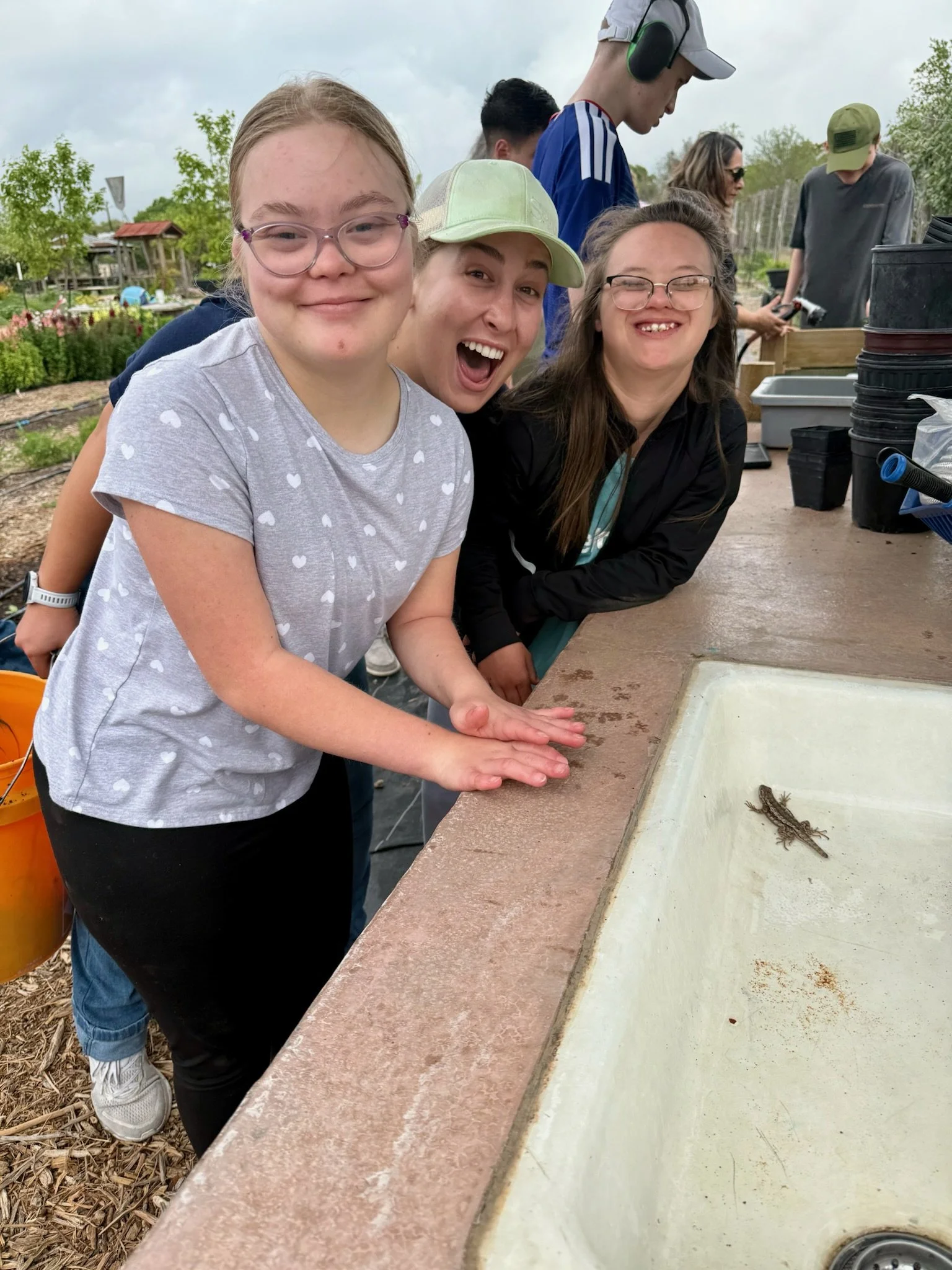Three children look at a small bug or vegetable at an outdoor educational area, with other people and trees in the background.