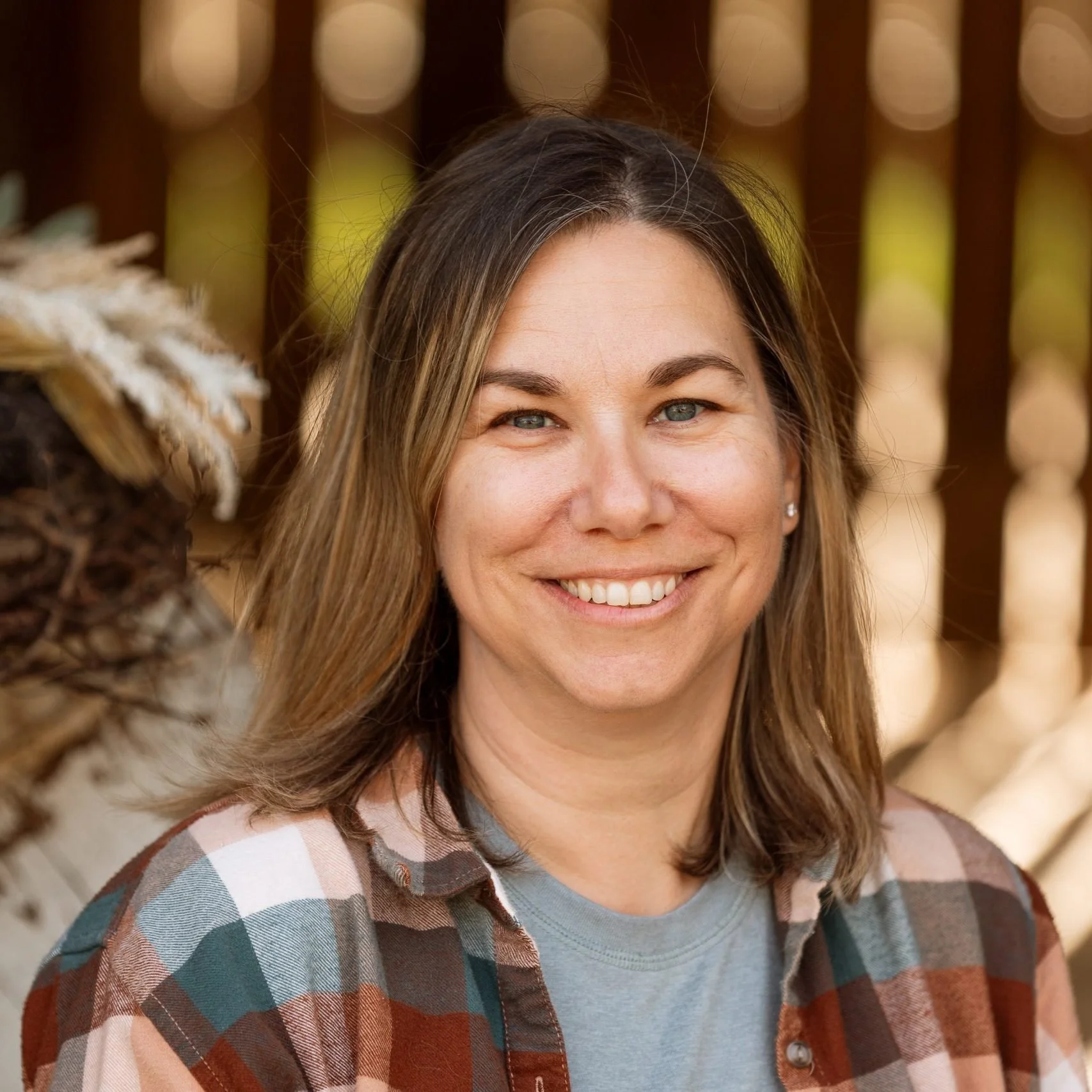 A smiling woman with blue eyes and brown hair, wearing a plaid shirt, standing outdoors with a blurred wooden fence and trees in the background.