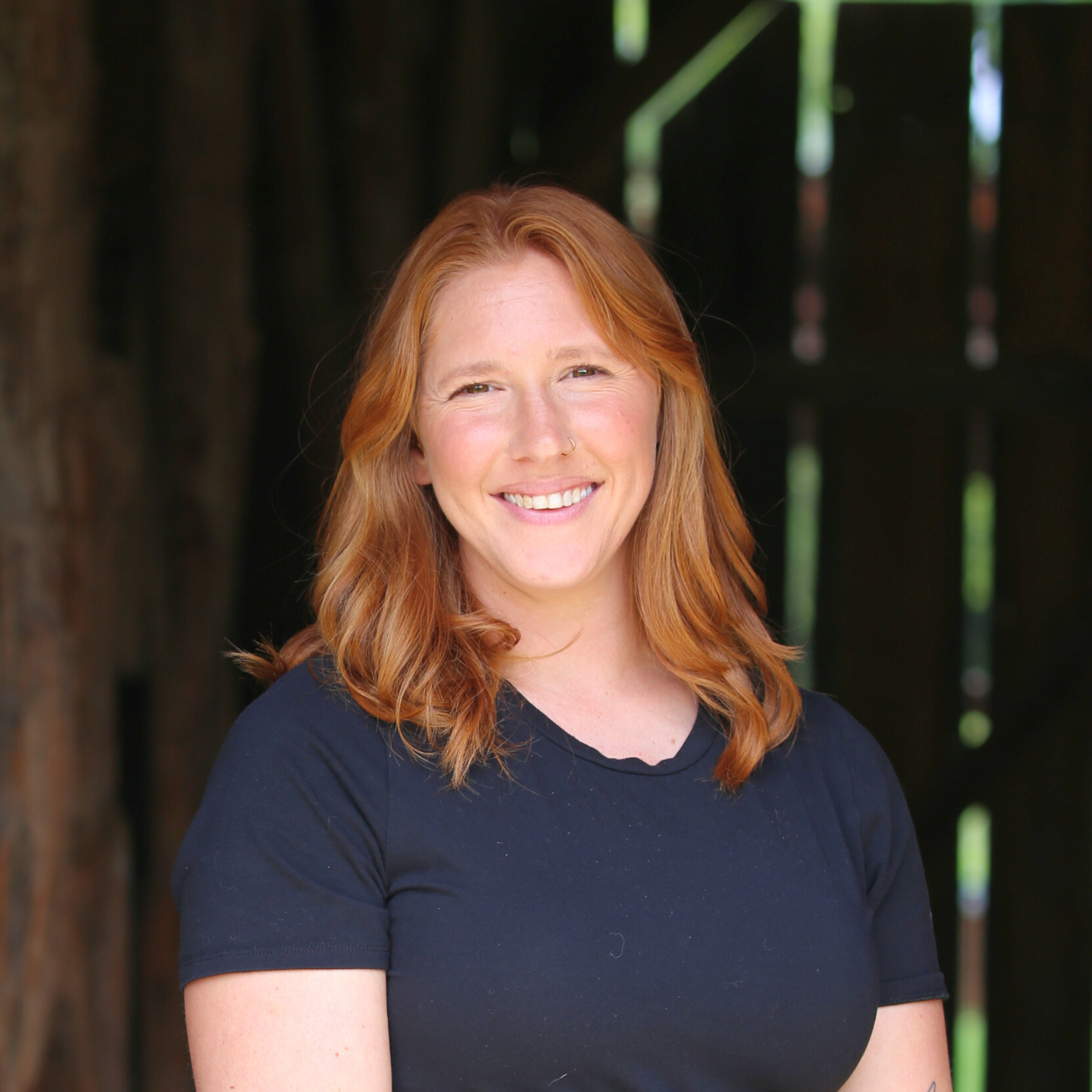 A young woman with red hair and a nose ring smiling outdoors in front of a dark wooded background.